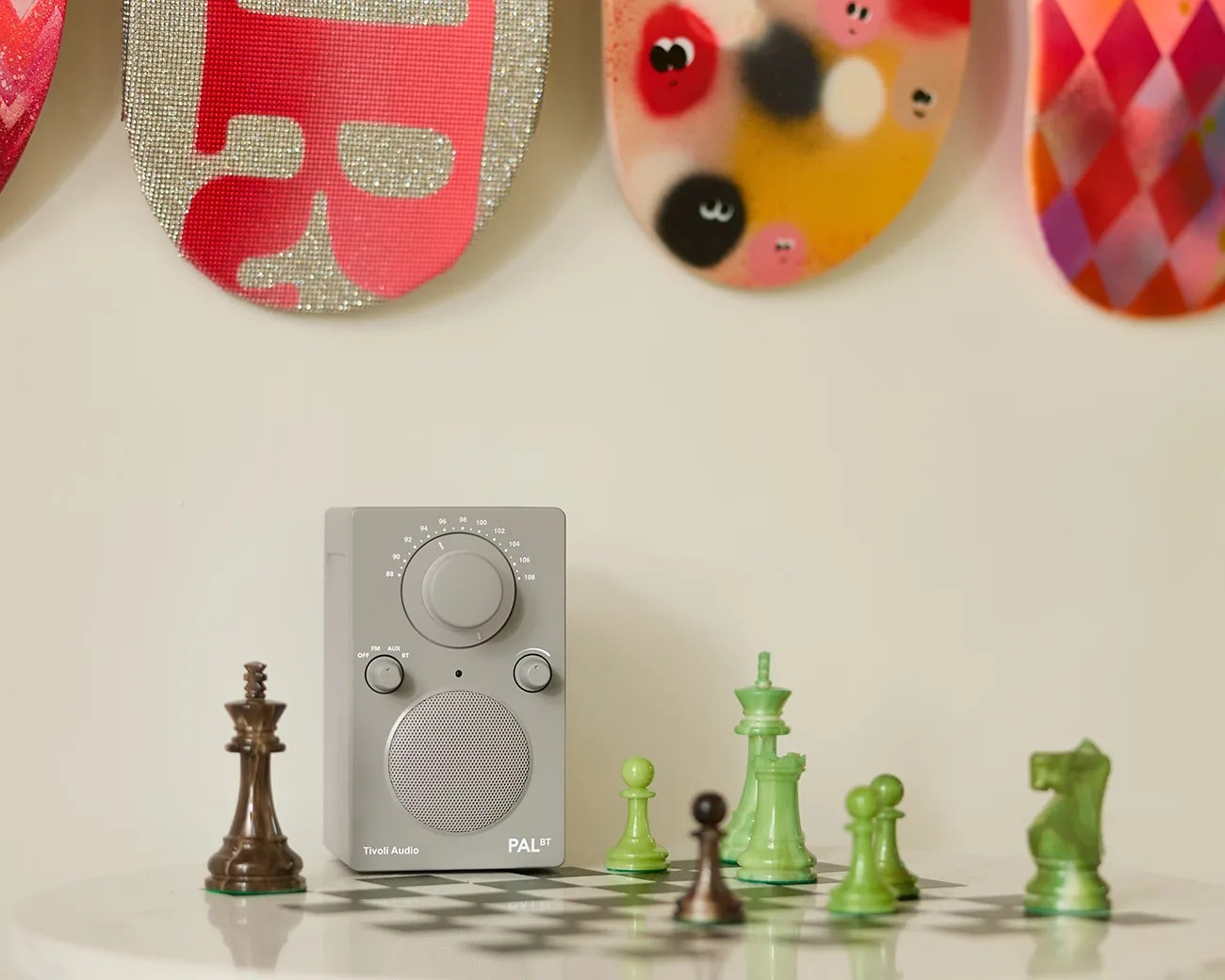 Gray Tivoli Audio PAL BT radio on a white table with green and brown chess pieces and colorful skateboard decks on the wall.