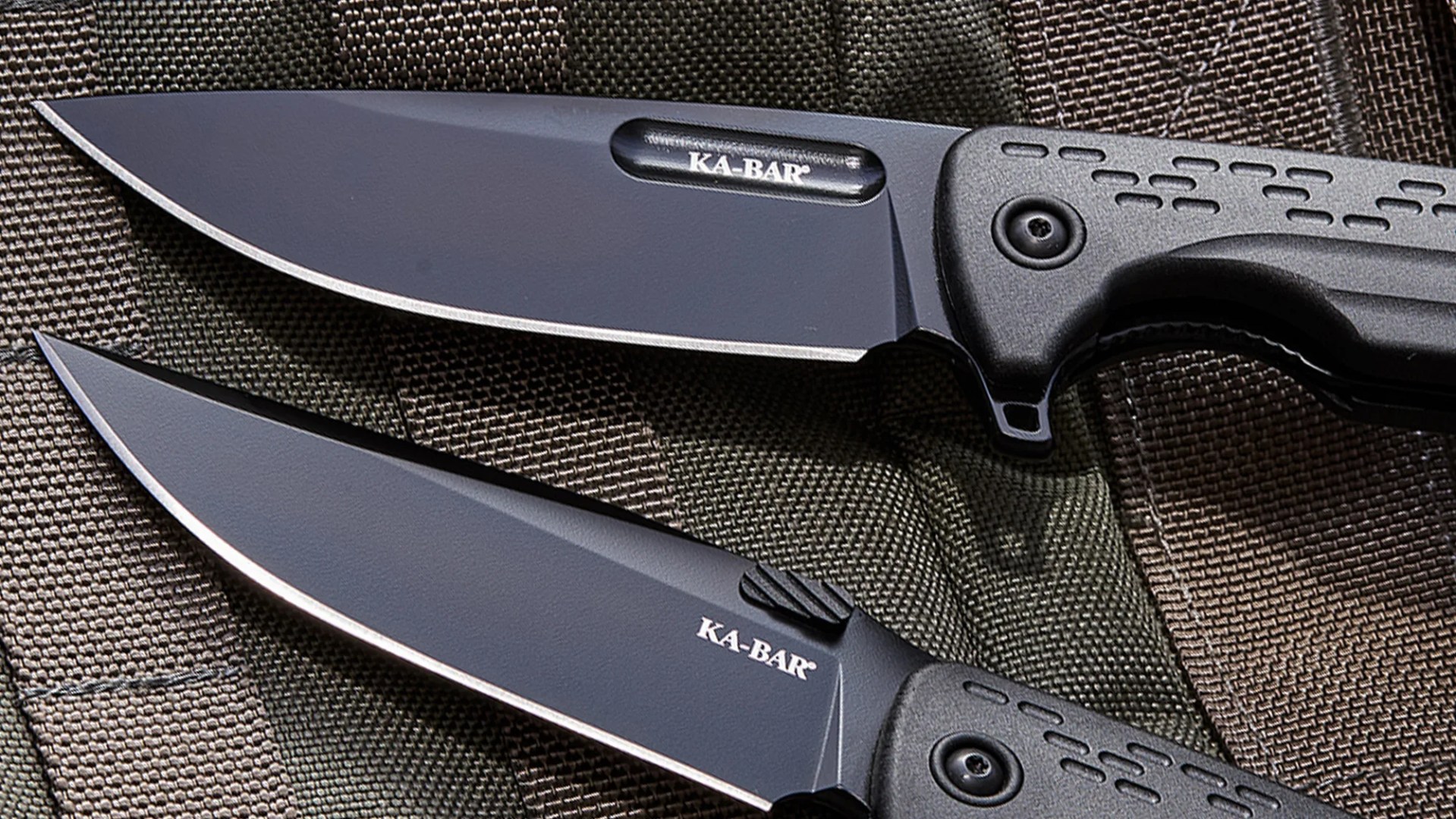 Two black KA-BAR folding knives with textured handles on a dark woven fabric surface.
