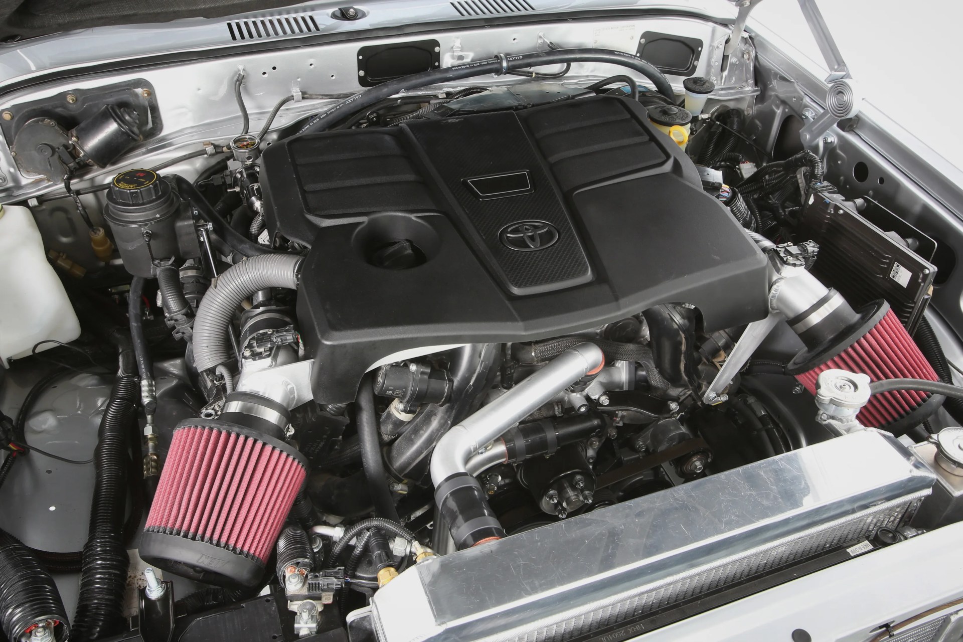 Engine bay with a black Toyota engine cover and dual red cone air filters connected by silver intake pipes.