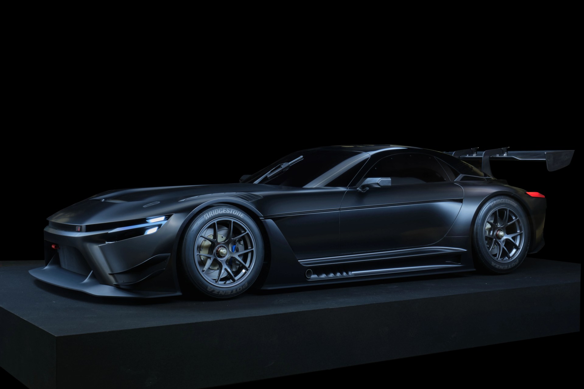 Matte black race car with aerodynamic body, large rear wing, and Bridgestone tires on a black platform.