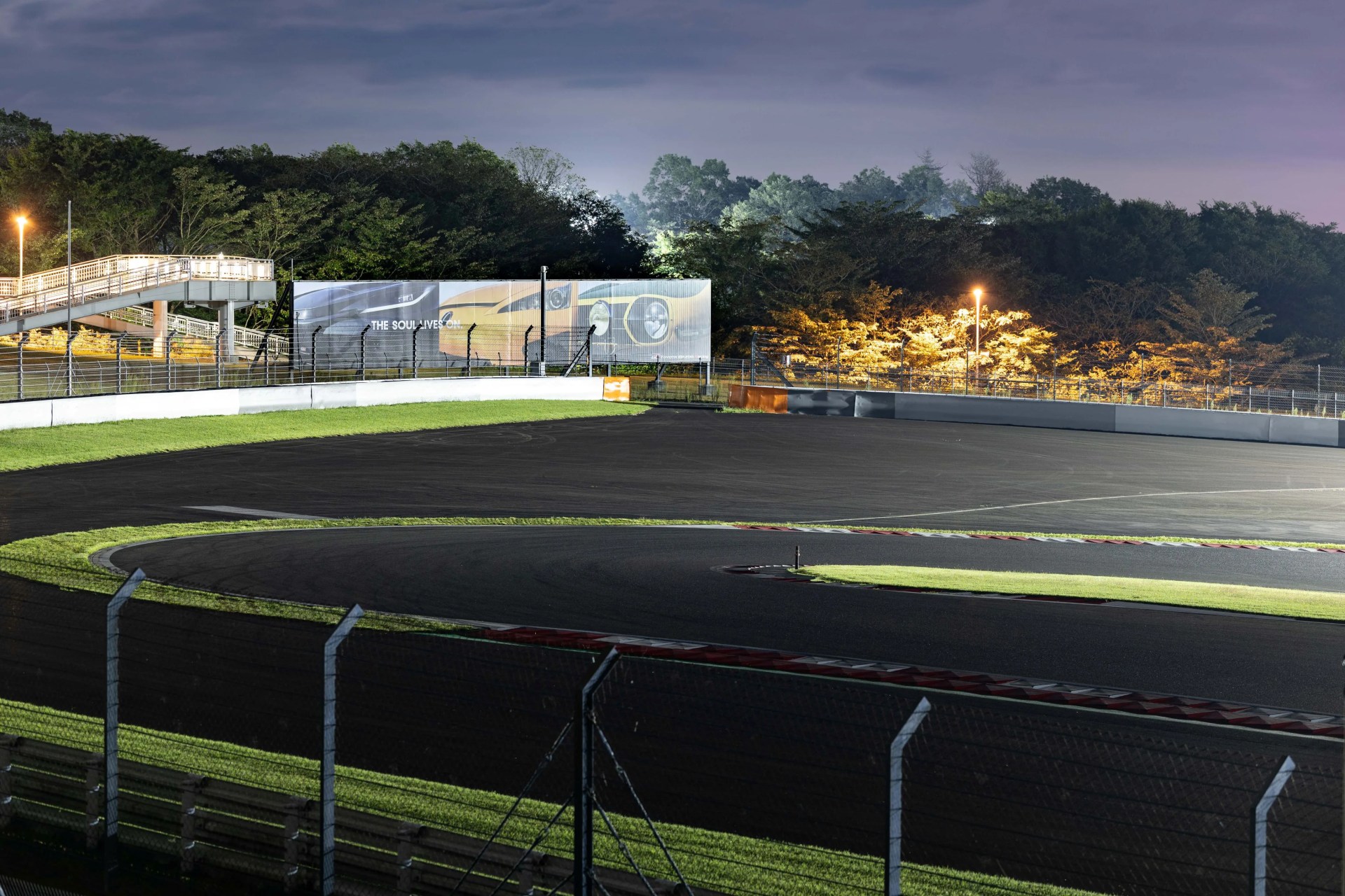 Night view of a curved race track with safety fences, illuminated trees, and a pedestrian bridge in the background.