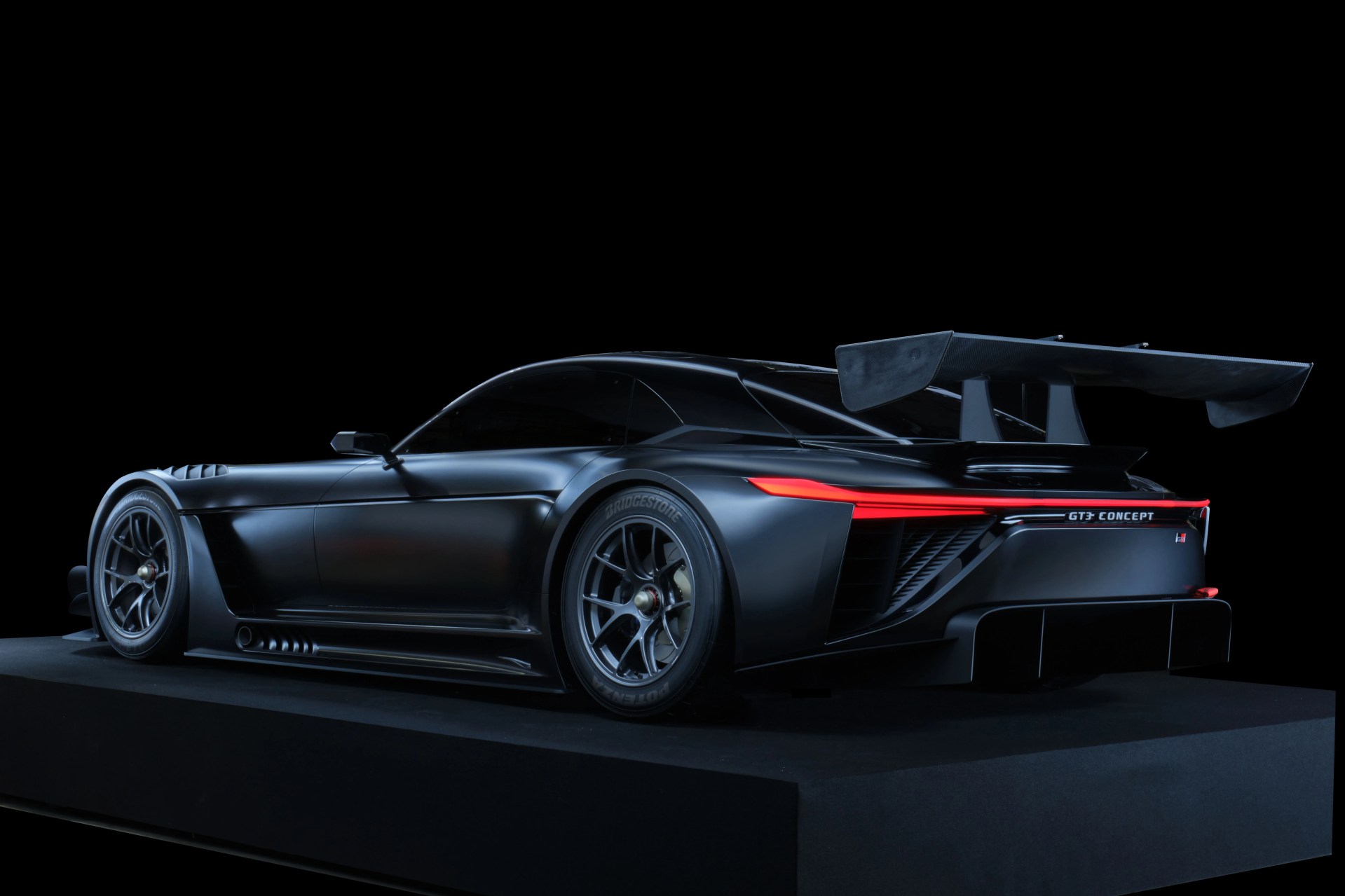 Matte black GT3 Concept race car with large rear wing and red LED taillights on a black platform.