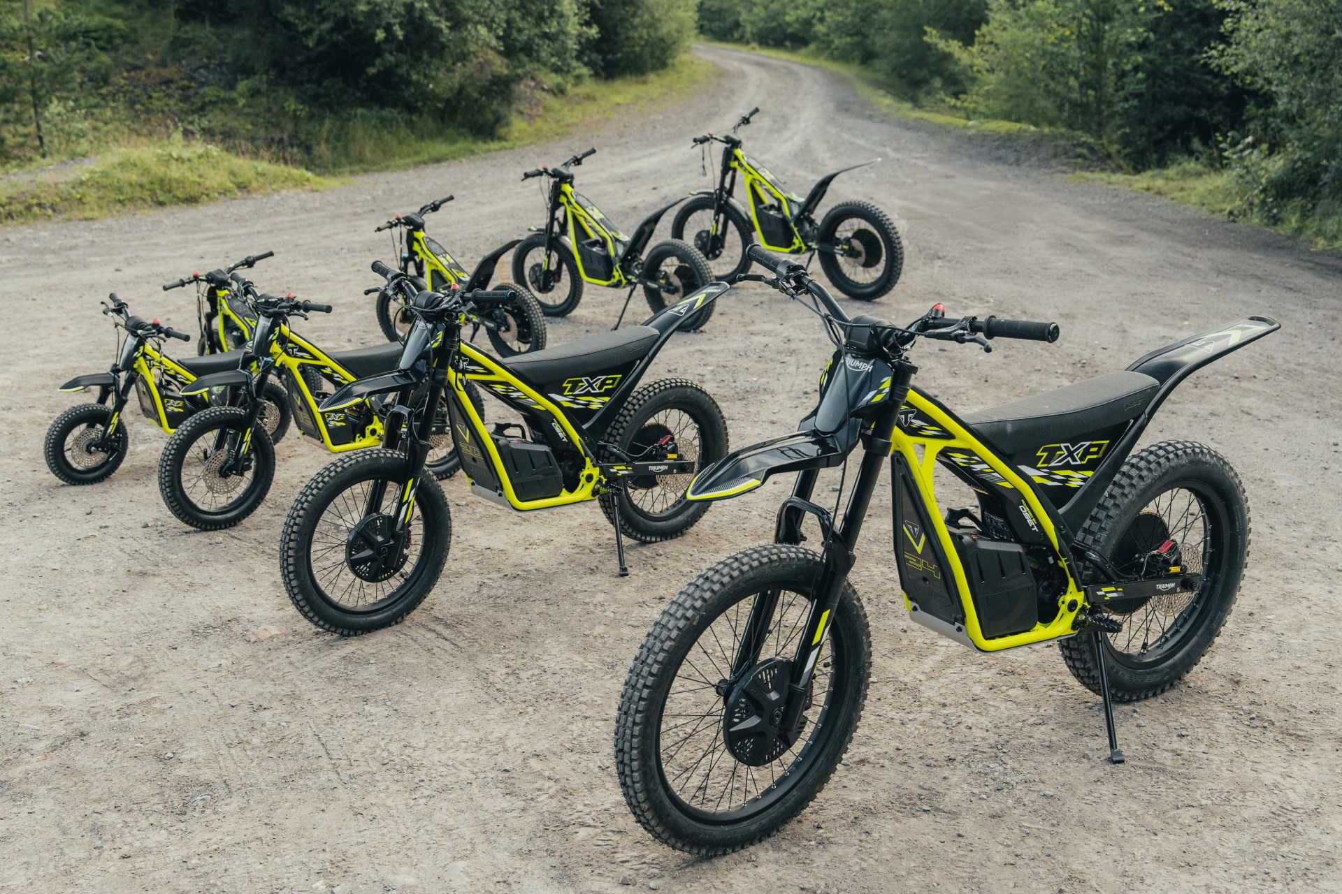 Seven black and neon yellow electric dirt bikes parked on a dirt path surrounded by greenery.