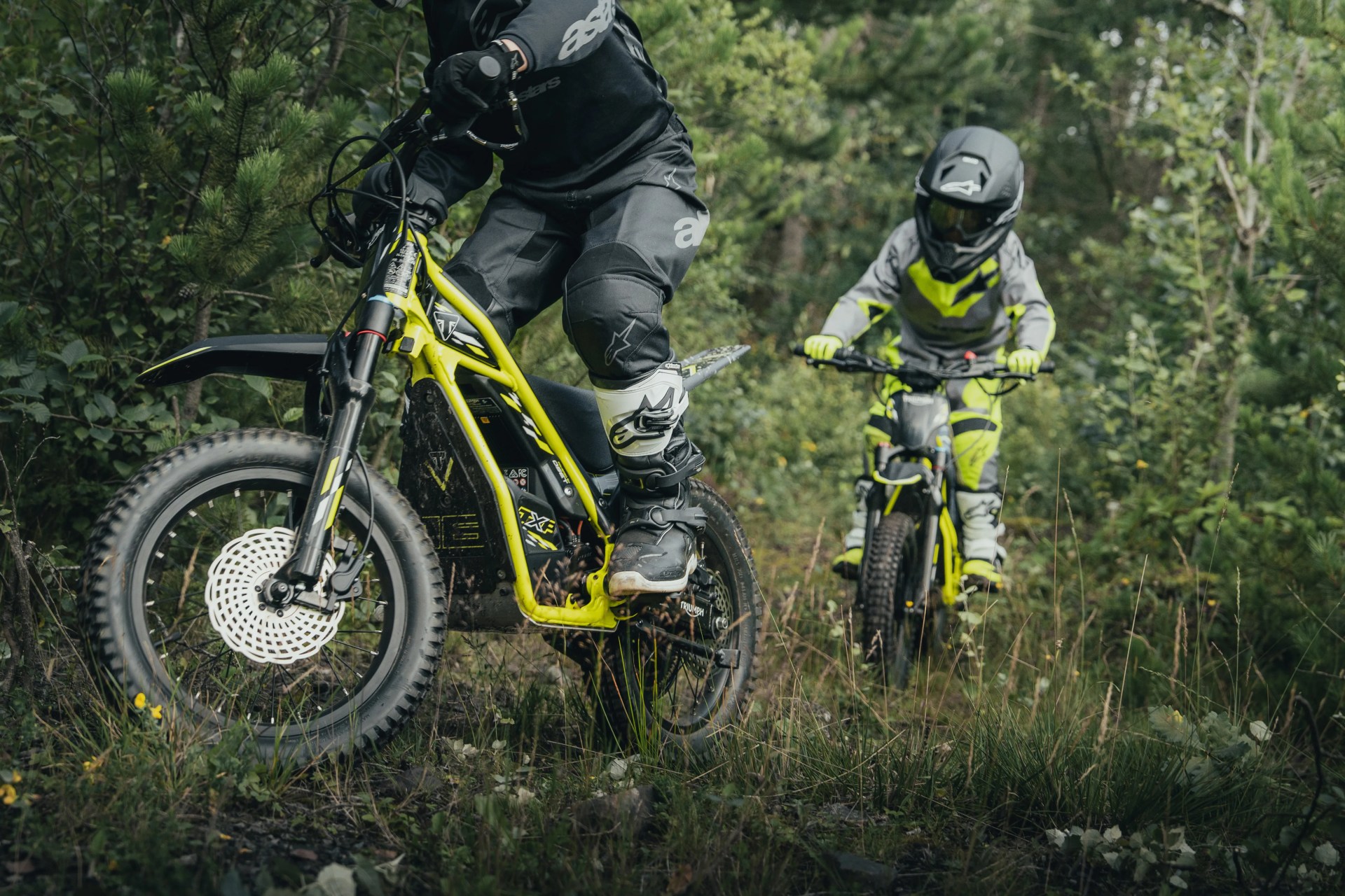 Two riders in black and neon yellow gear riding yellow electric dirt bikes on a forest trail.