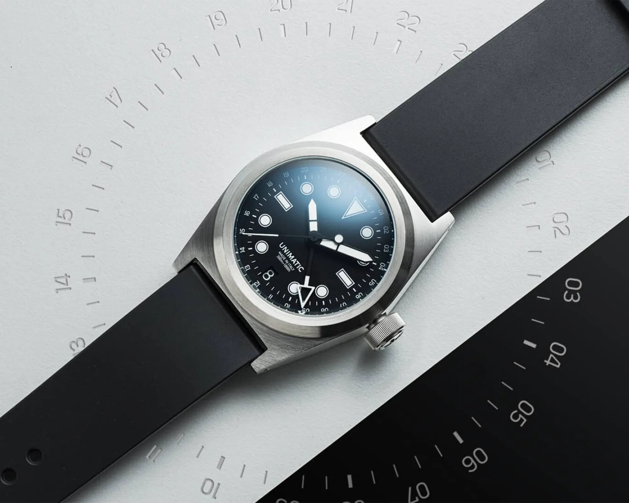 Stainless steel wristwatch with black dial, white markers, and black rubber strap on a white and black background.