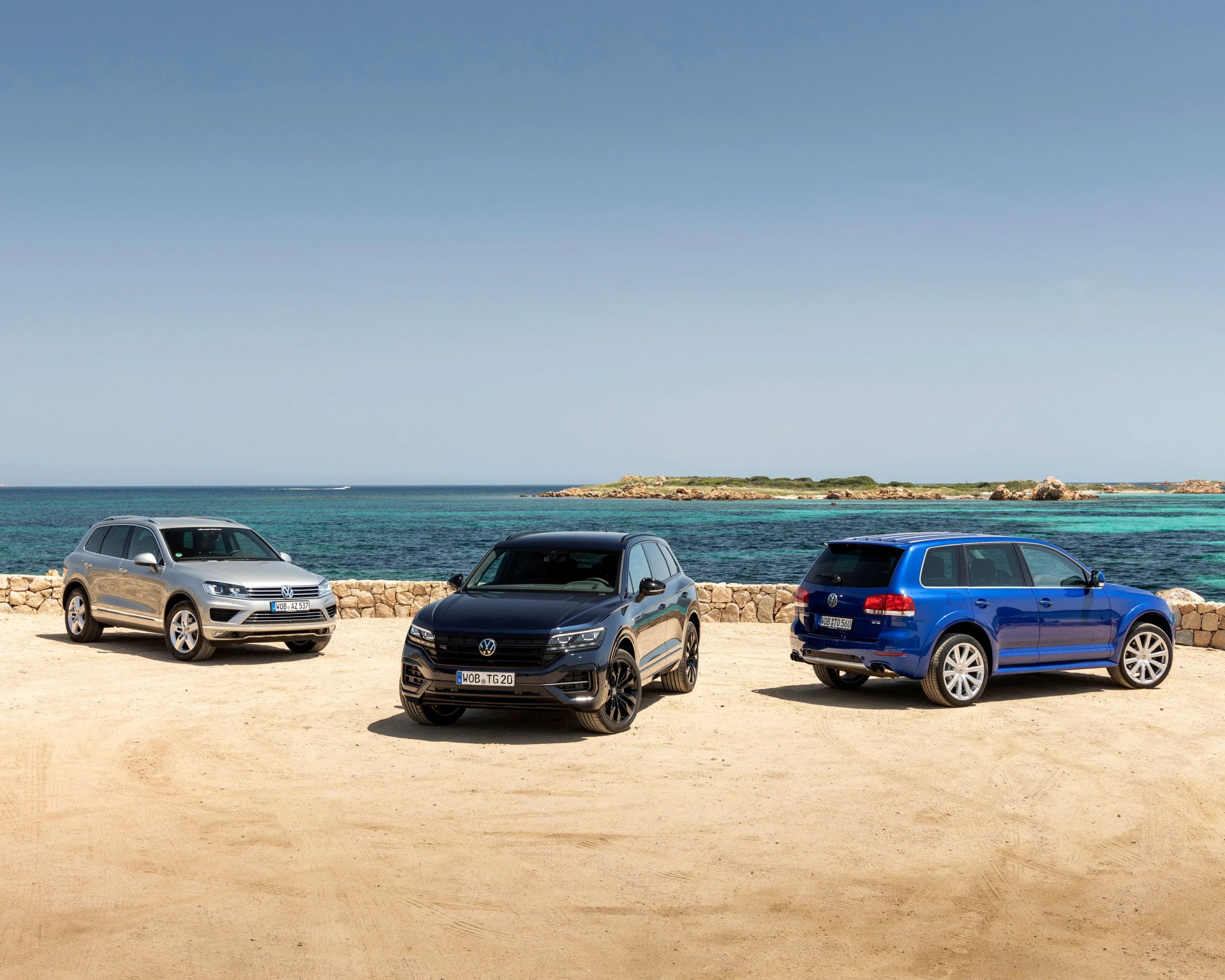 Three Volkswagen SUVs in silver, black, and blue parked on sandy ground by a stone wall with turquoise sea and clear sky in the background.