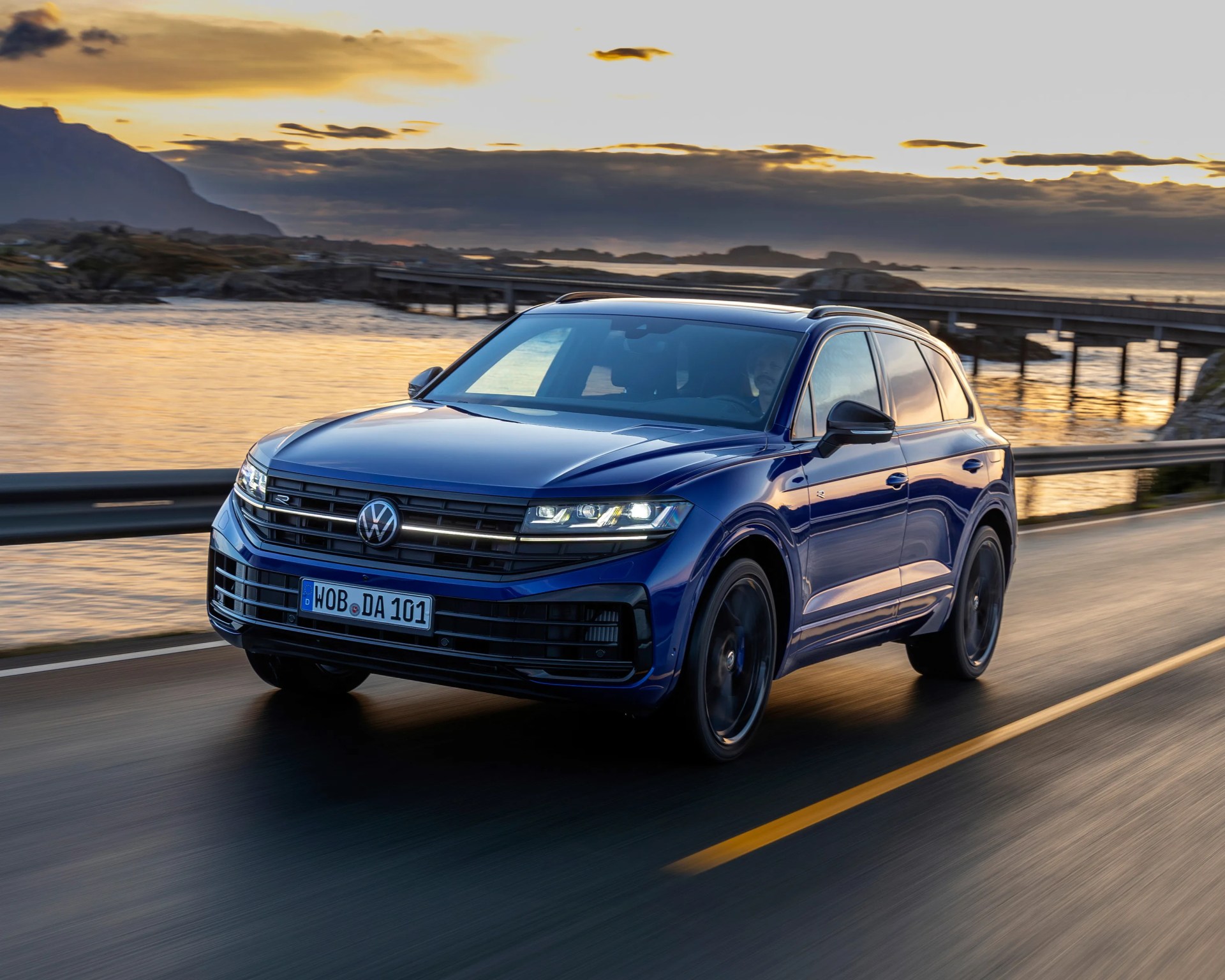 Blue Volkswagen SUV driving on a coastal road at sunset with water and mountains in the background.