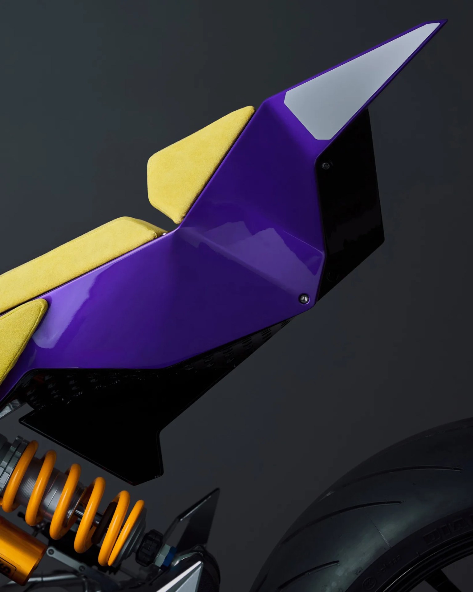 Close-up of a motorcycle seat with yellow cushions and a glossy purple and black angular body panel above an orange coil suspension and rear tire.