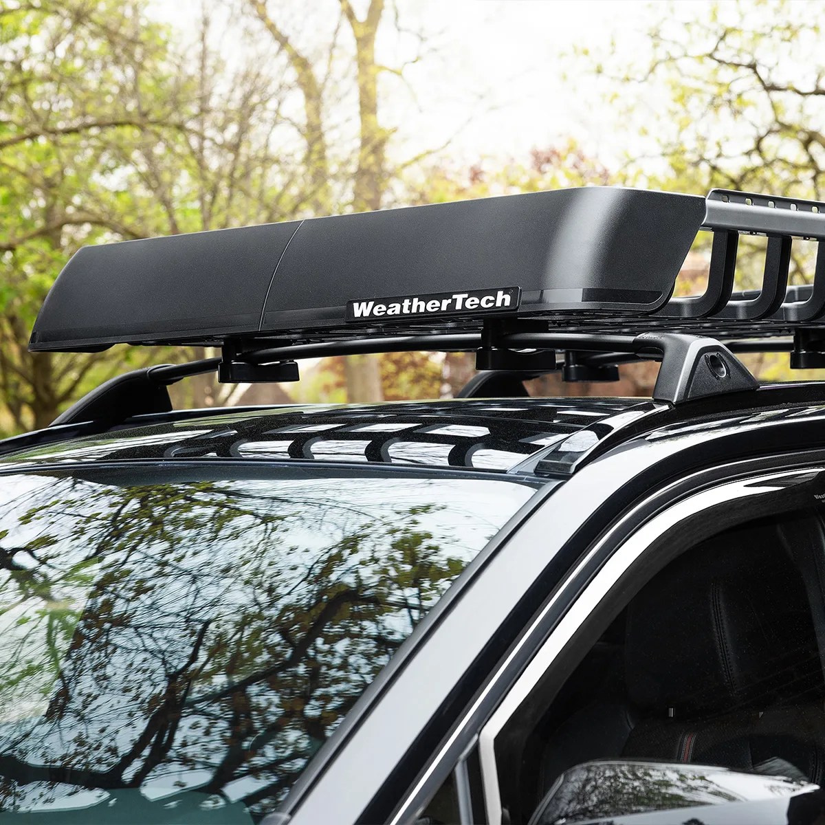 Black WeatherTech roof cargo basket mounted on a black vehicle with trees reflected on the windshield.