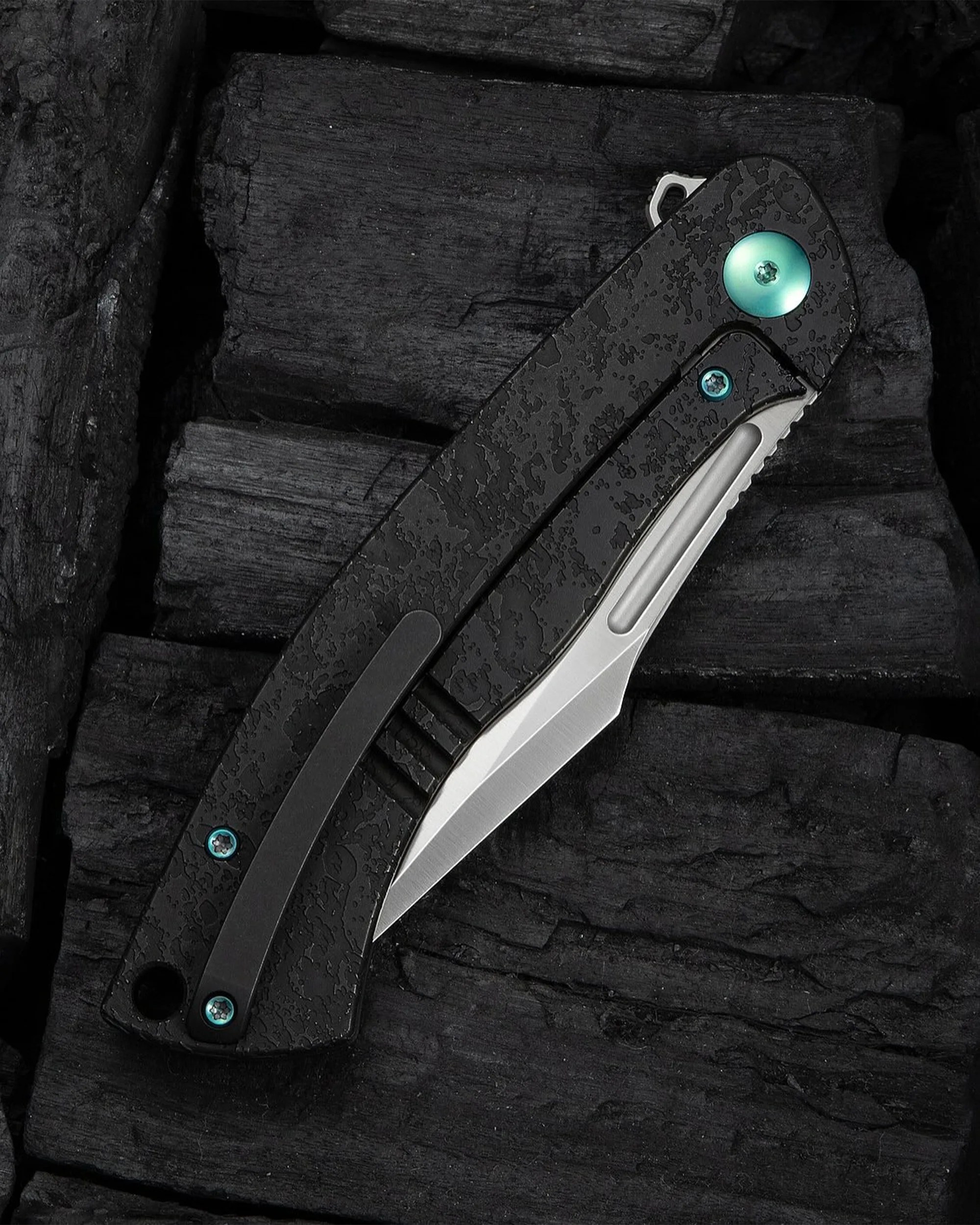 Black textured folding knife with partially exposed silver blade and turquoise screws on dark charcoal background.