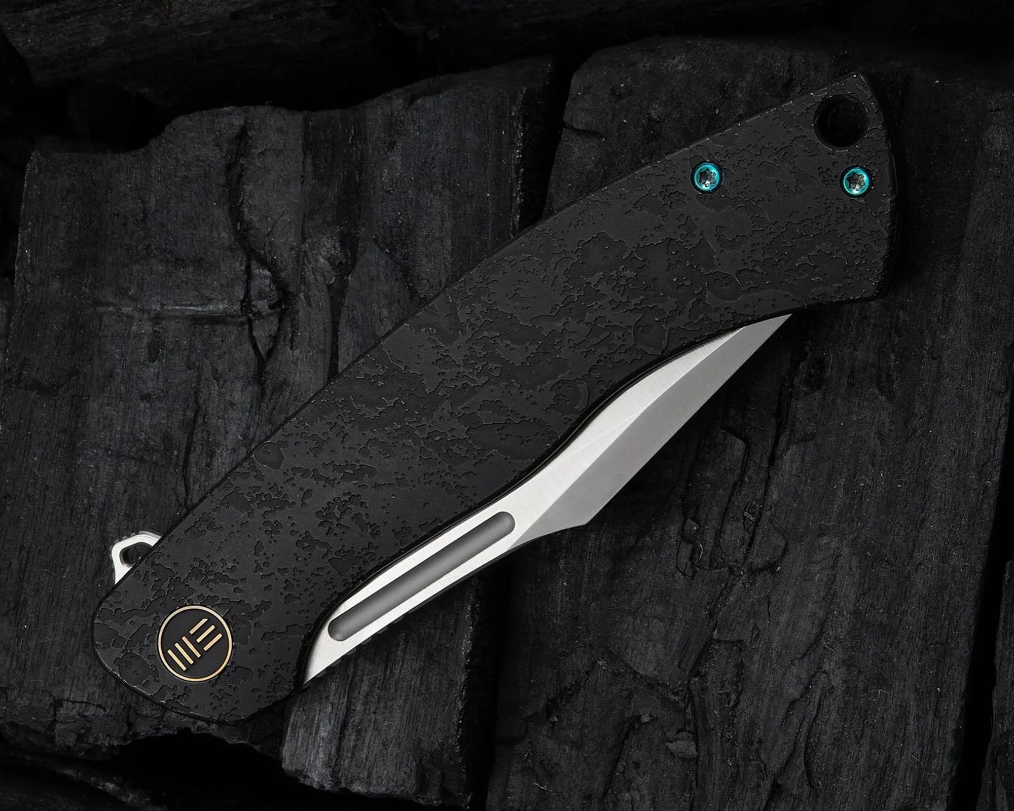 Black textured folding knife with a partially visible silver blade and blue screws on a dark wooden surface.