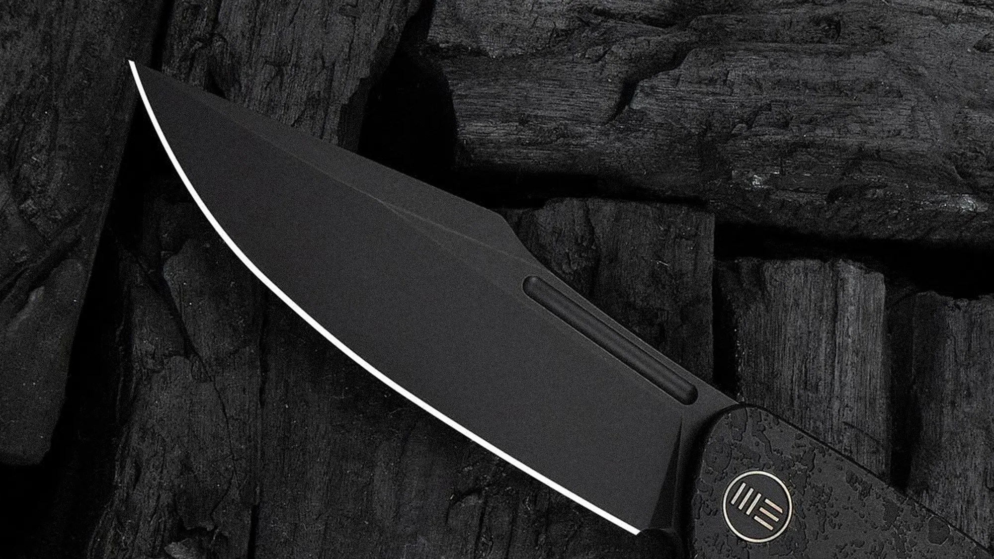 Black folding knife blade with a matte finish and a textured handle on dark wooden surface.