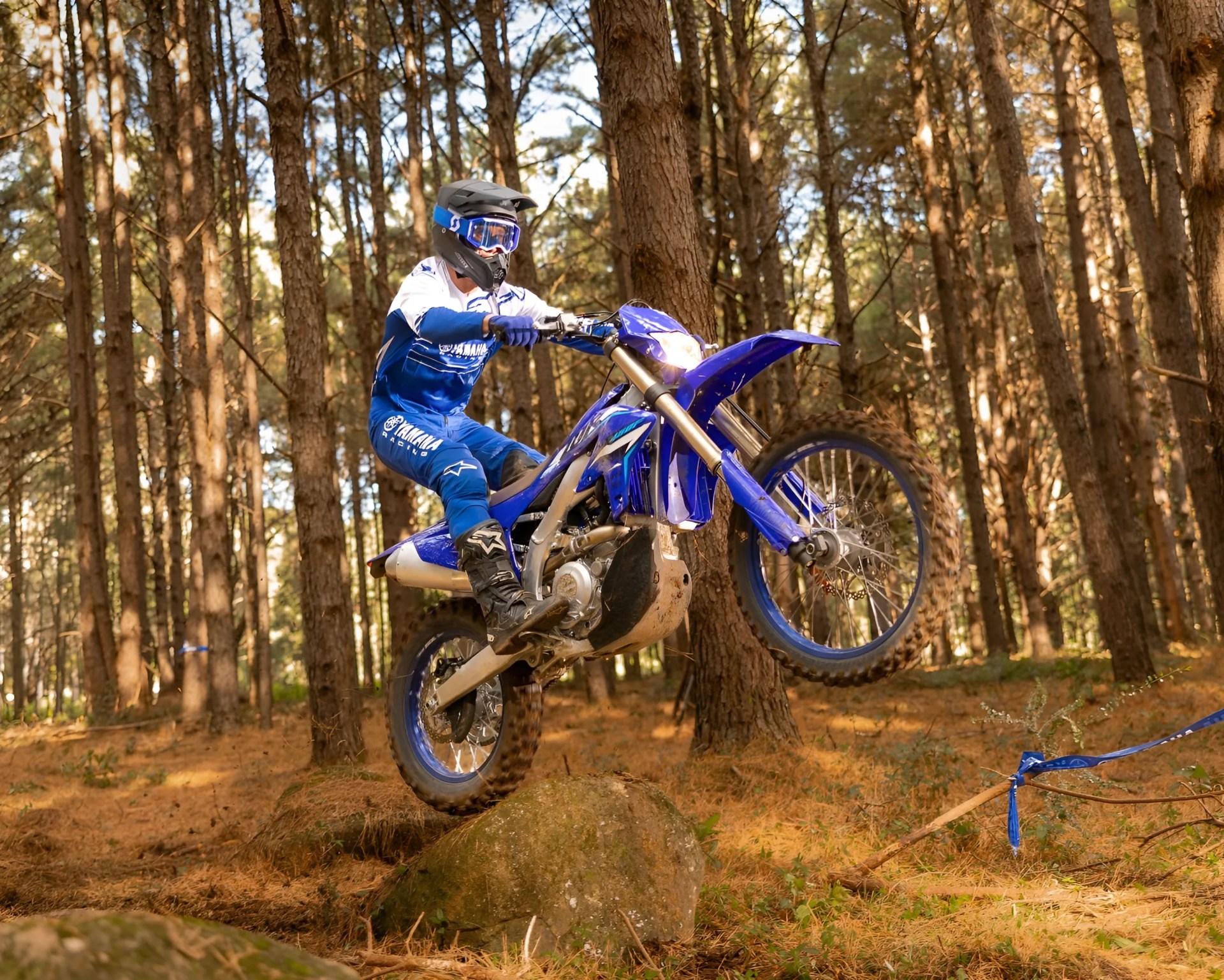 Person in blue and white Yamaha racing gear riding a blue dirt bike over a rock in a forest.