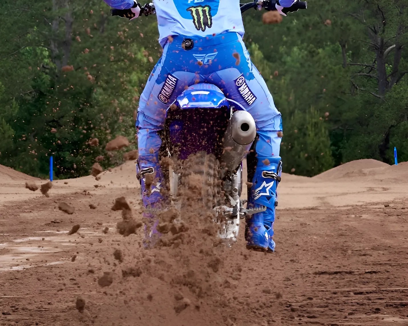 Rear view of a person in blue Yamaha motocross gear riding a dirt bike on a muddy track with dirt flying up.