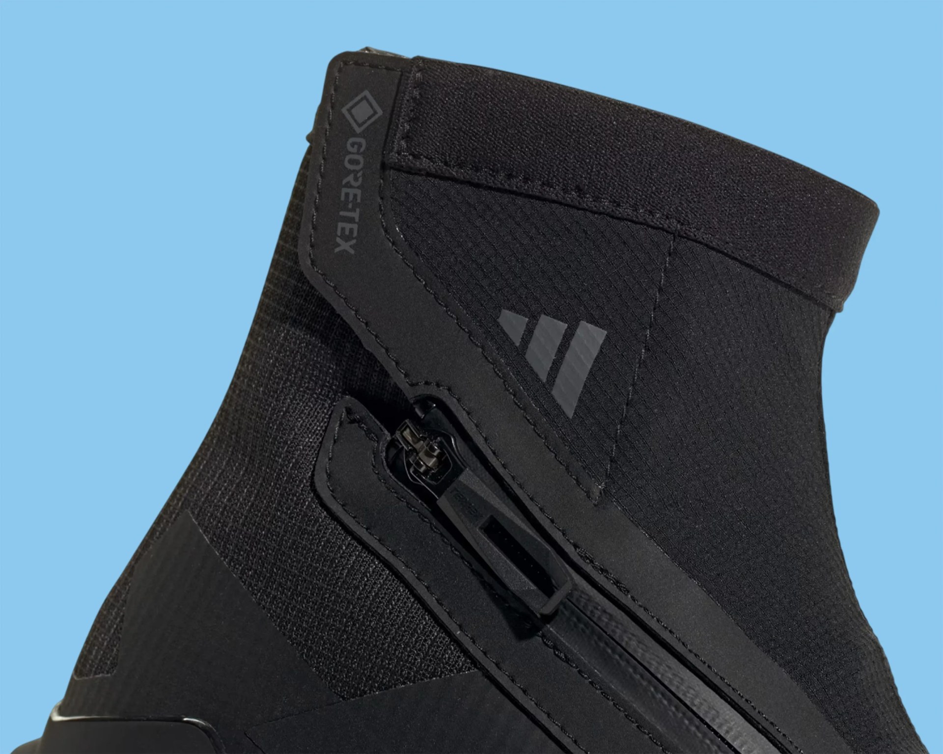 Close-up of black Gore-Tex fabric shoe upper with zipper and Adidas logo on a blue background.