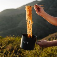 Black camping pot with noodles held by chopsticks and a hand outdoors with hills in the background.