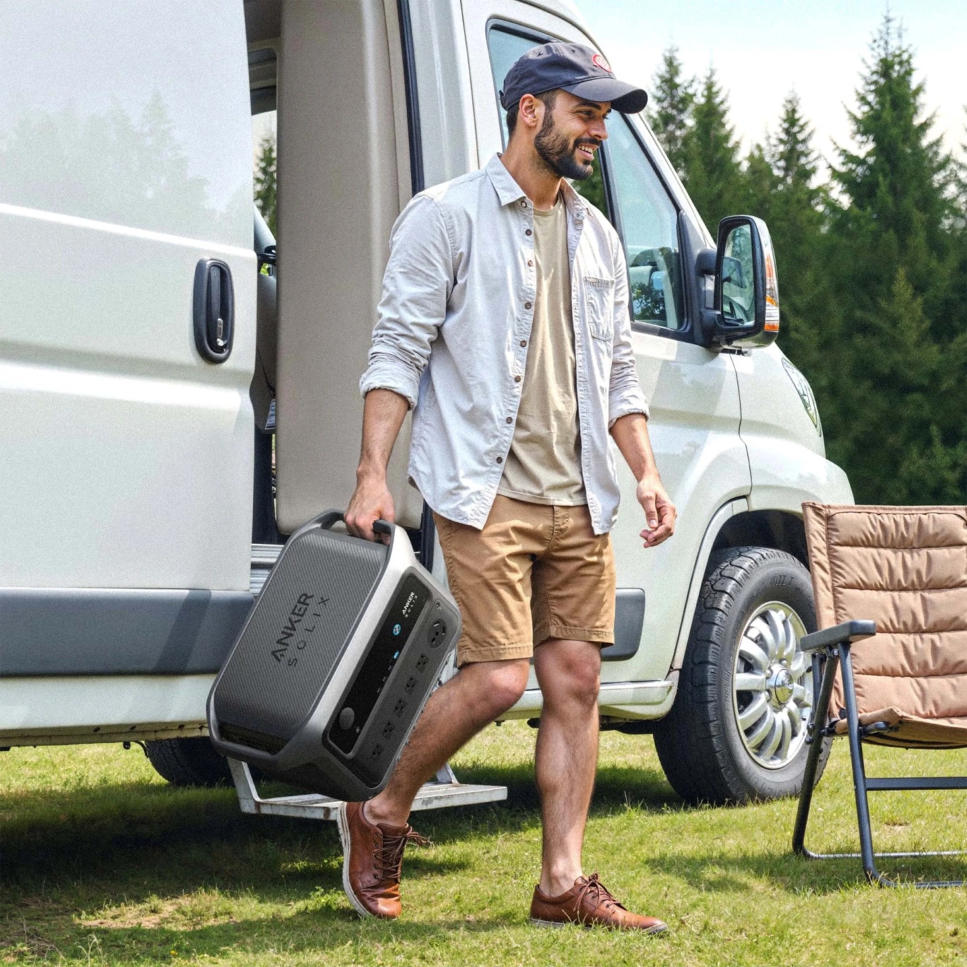 Man carrying a gray Anker Solix portable power station near a white van and camping chair on grass.