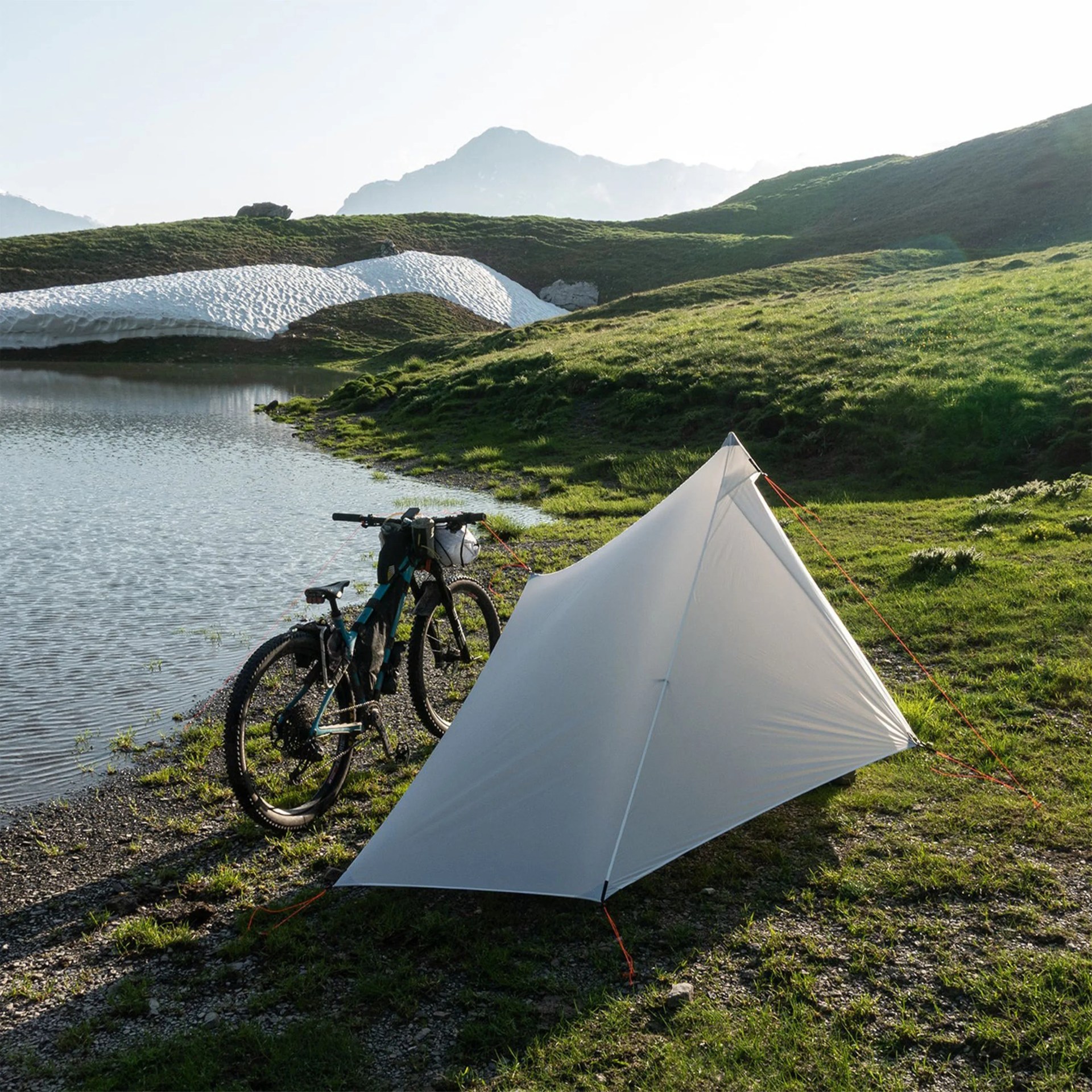 Light gray triangular tent pitched on grassy ground near a lake with a loaded mountain bike beside it.