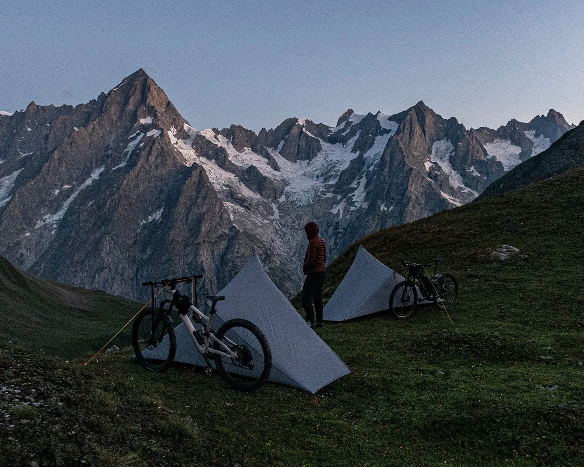 Two gray tents pitched on grassy hillside with two mountain bikes and a person in a red jacket, snowy rocky peaks in background.