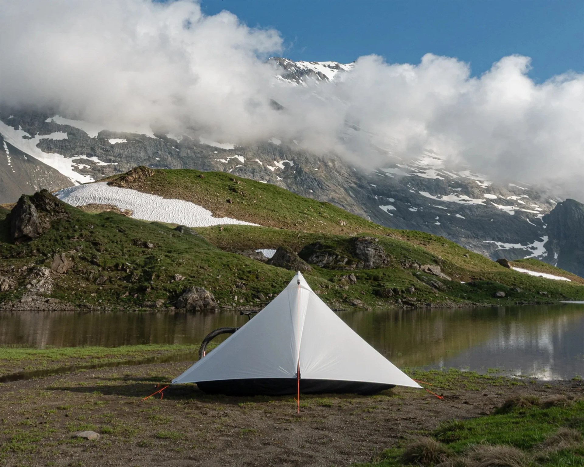 White triangular tent pitched on grassy ground near a mountain lake with snow patches and clouds overhead.