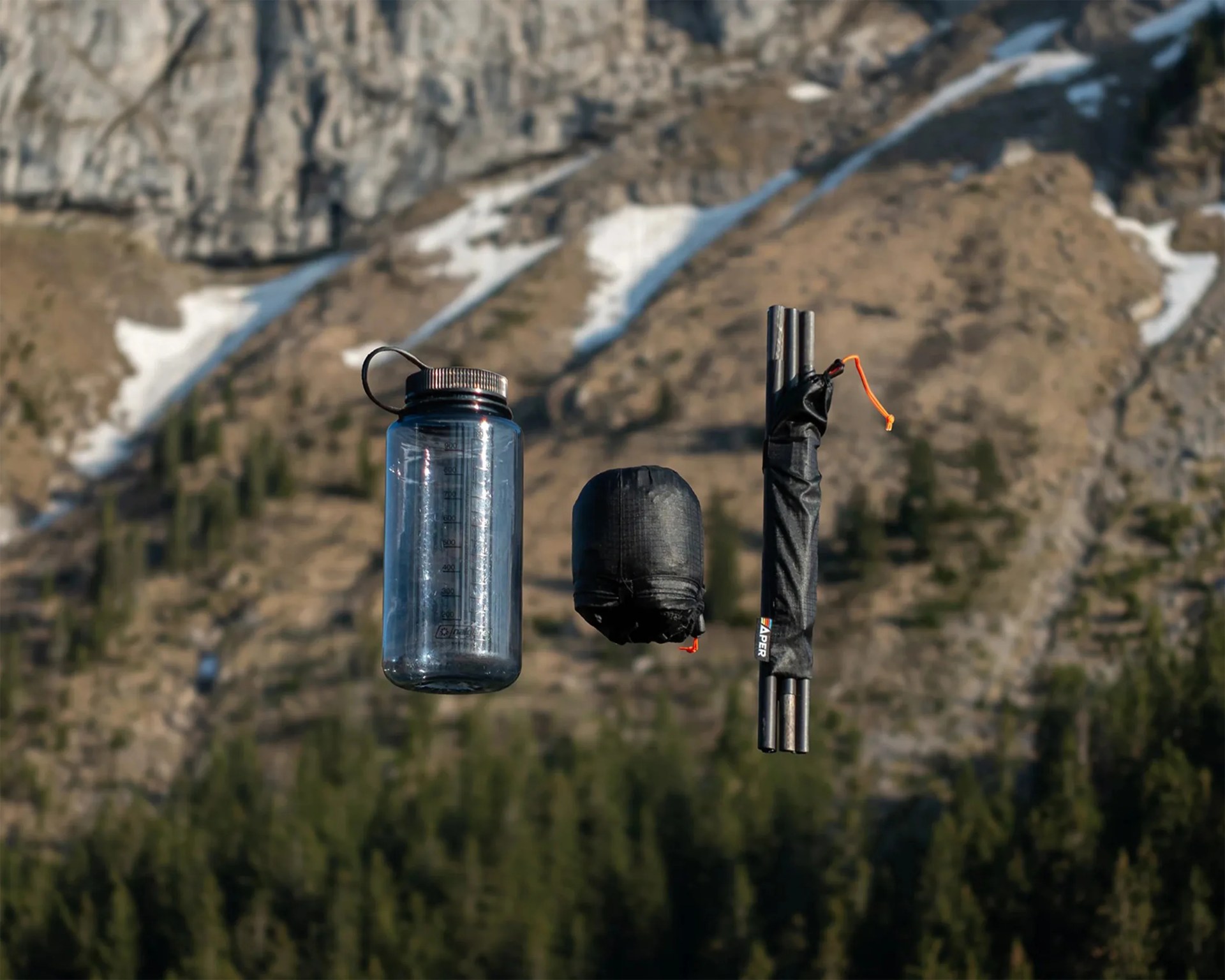 Clear plastic water bottle, black stuff sack, and folded black trekking poles with orange cord, set against a mountainous background.