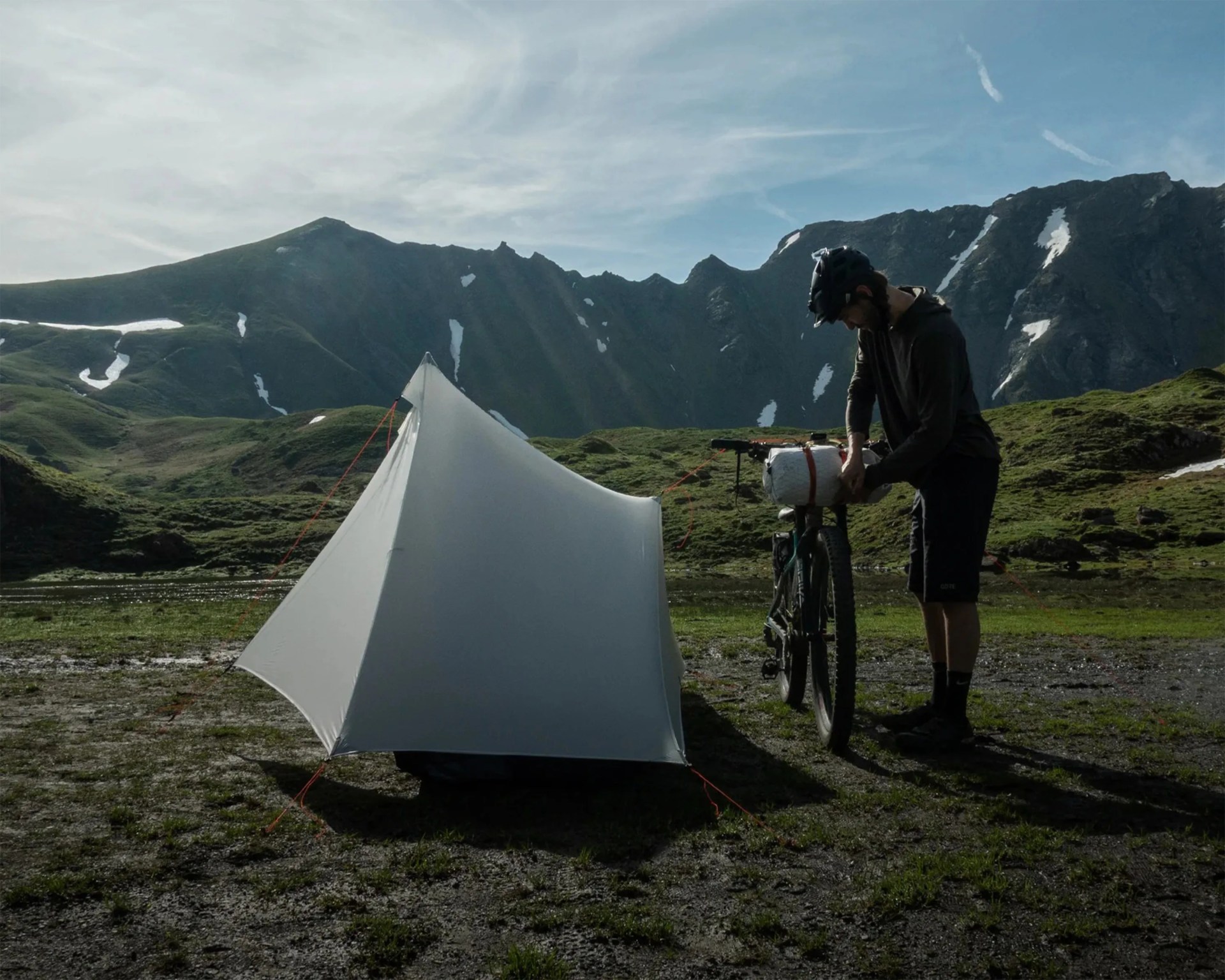 White triangular tent pitched on grassy ground with a person securing gear on a mountain bike in a mountainous landscape.