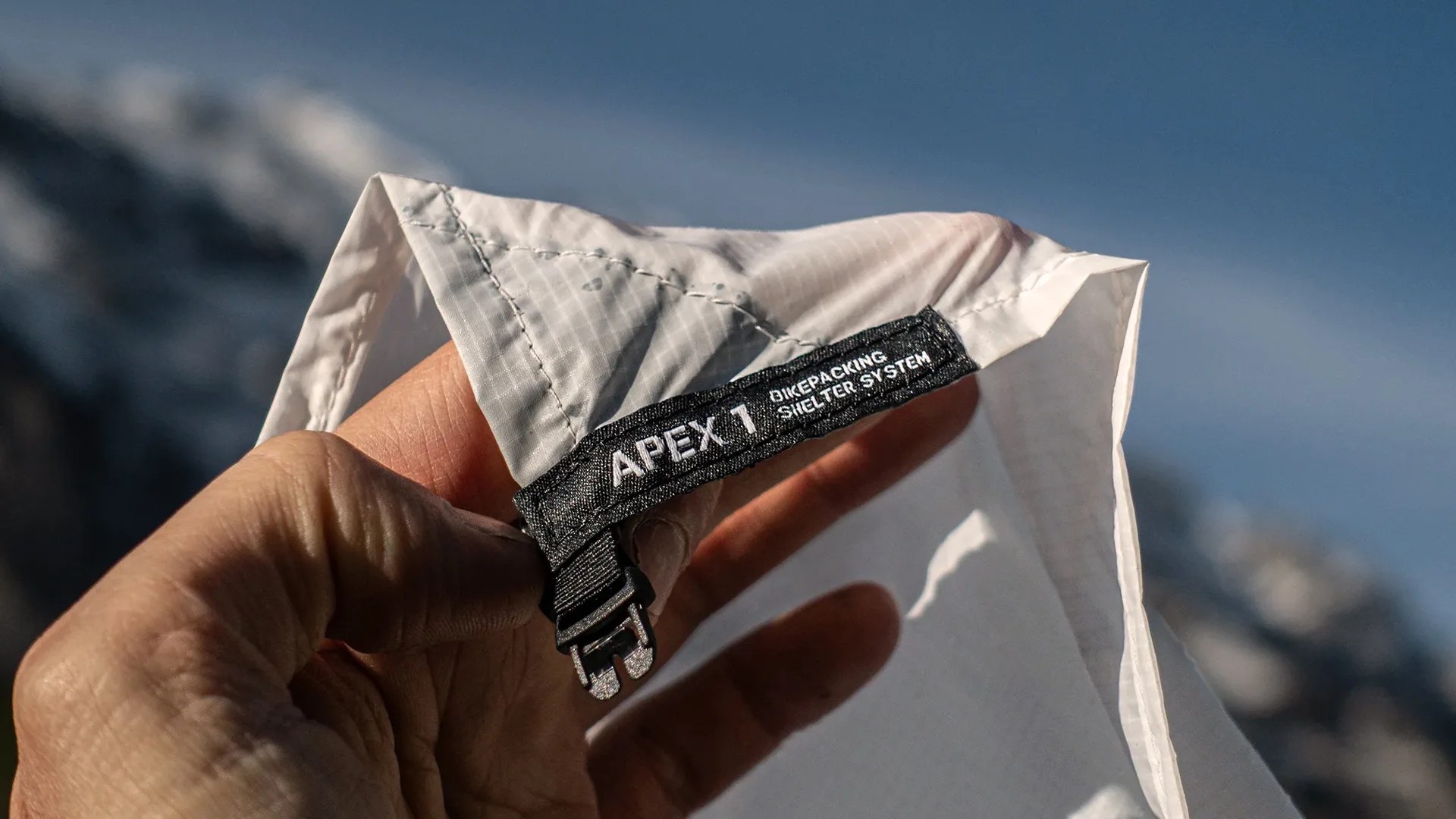 Hand holding a white fabric corner with a black label reading "APEX 1 BIKEPACKING SHELTER SYSTEM" and a small buckle.