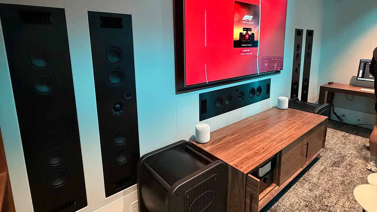 Living room entertainment setup with a large flat-screen TV, built-in wall speakers, two white smart speakers on a wooden cabinet, and a subwoofer on the floor.