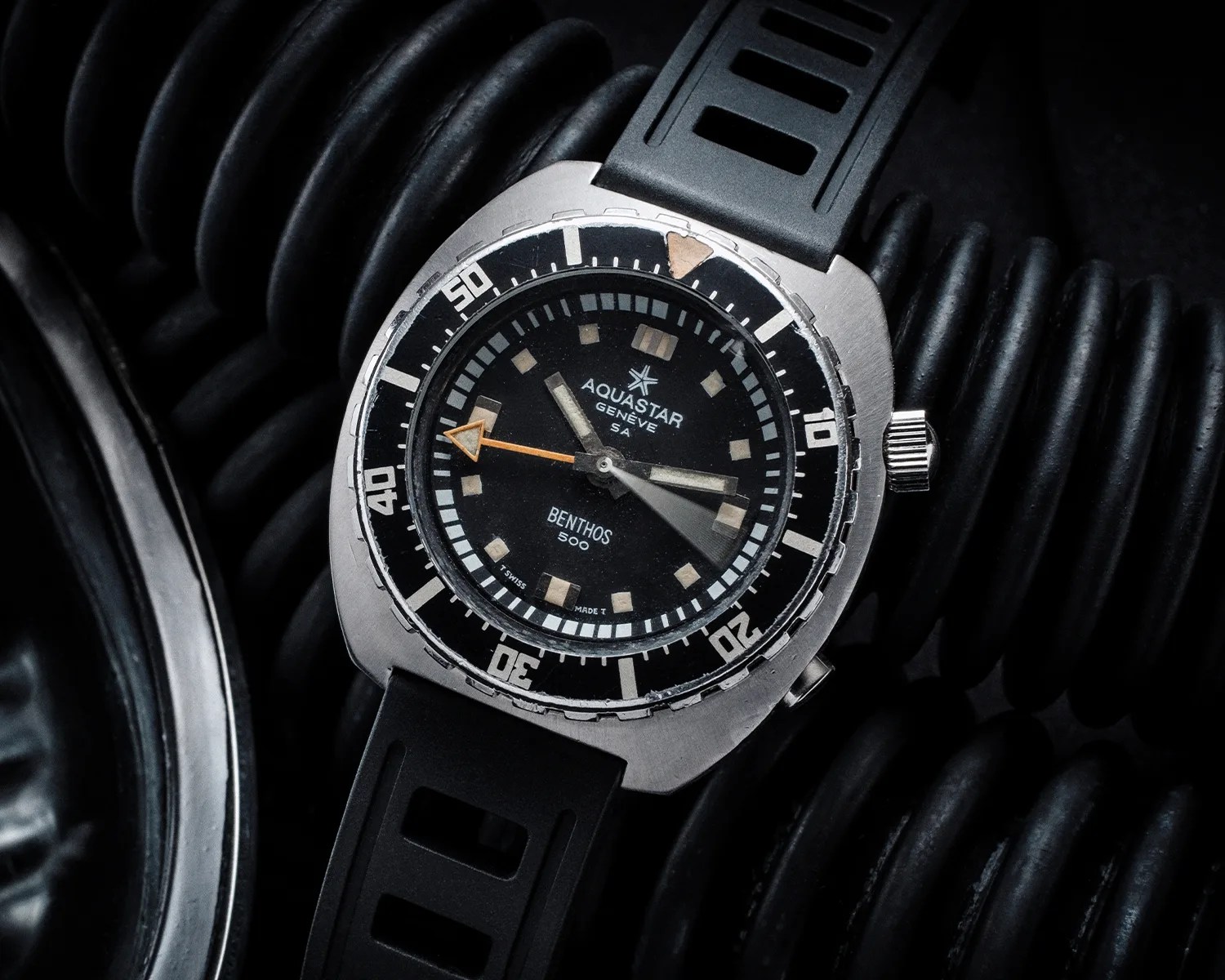 Vintage Aquastar Benthos 500 dive watch with black dial, orange arrow second hand, and black rubber strap on coiled hose.