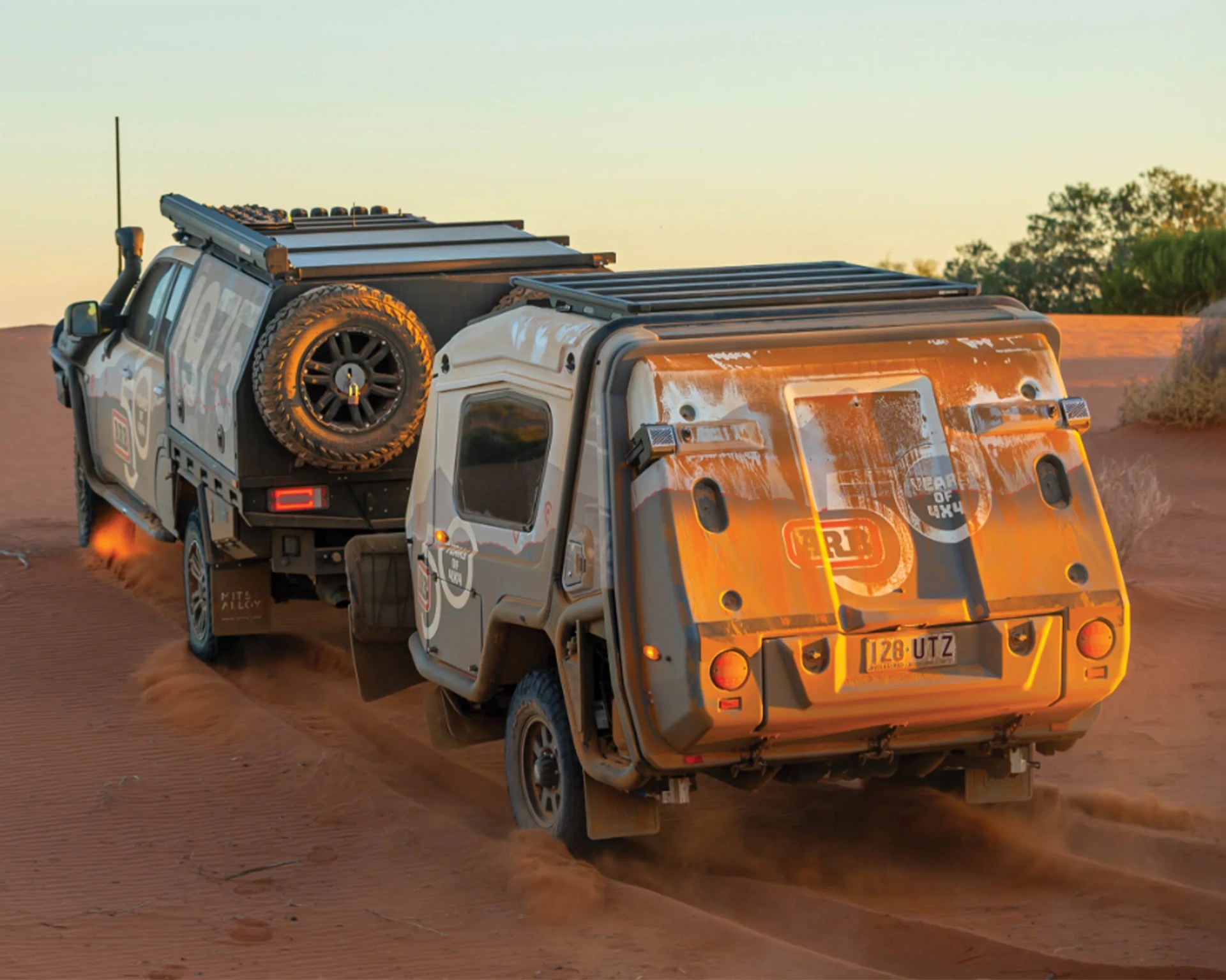 Off-road vehicle towing a rugged camper trailer on sandy terrain at sunset.