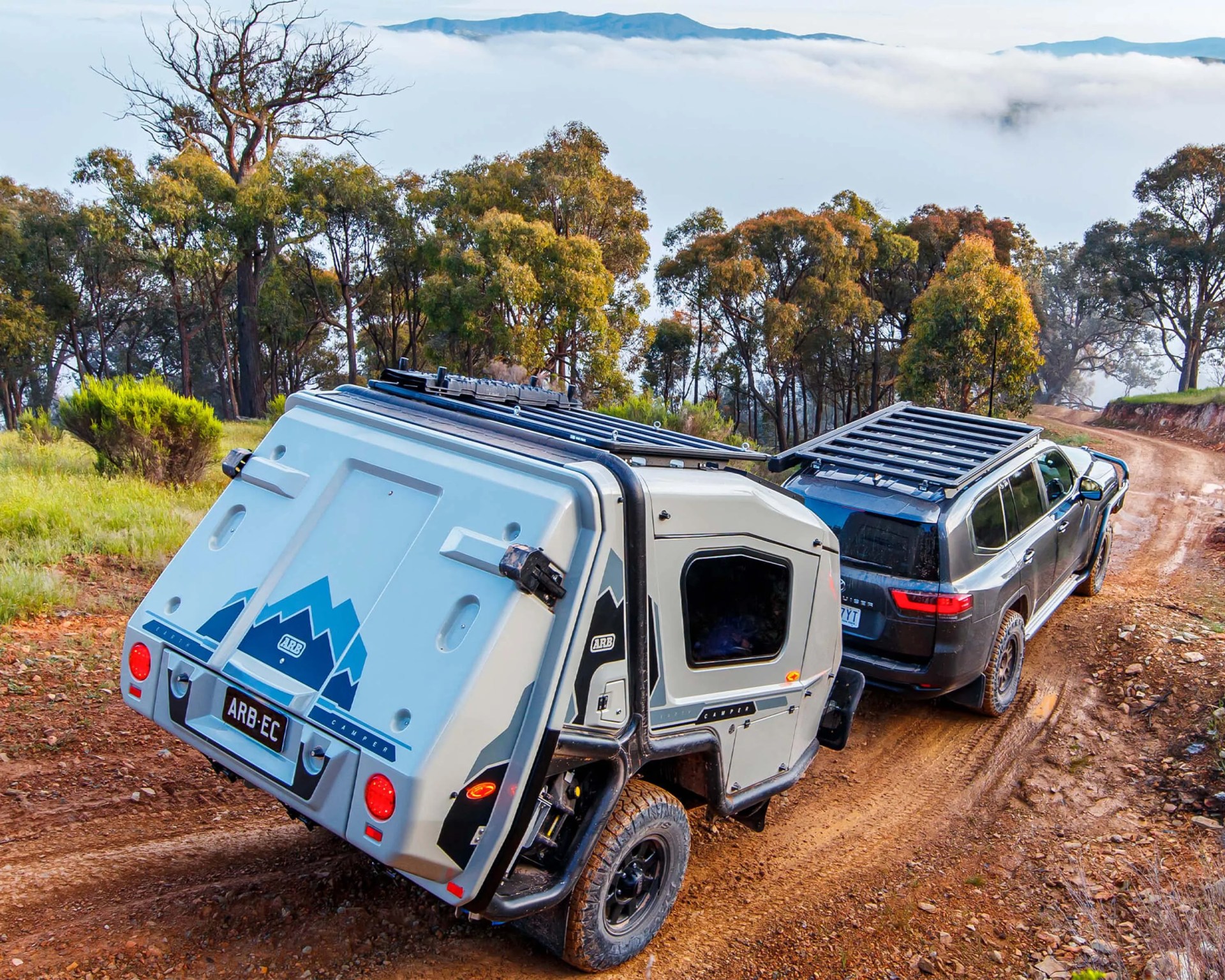Gray off-road camper trailer with mountain logo hitched to a black SUV on a dirt road in a forested area.