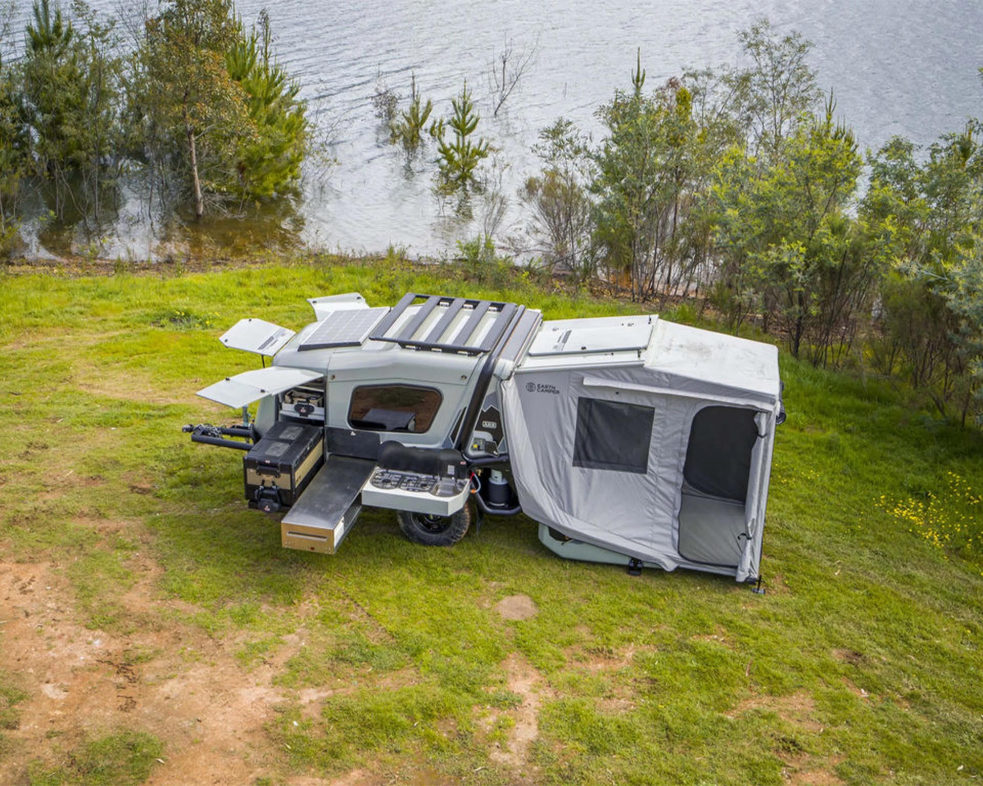 Compact off-road camper trailer with pop-up tent, solar panel, and outdoor kitchen setup on grassy lakeside.