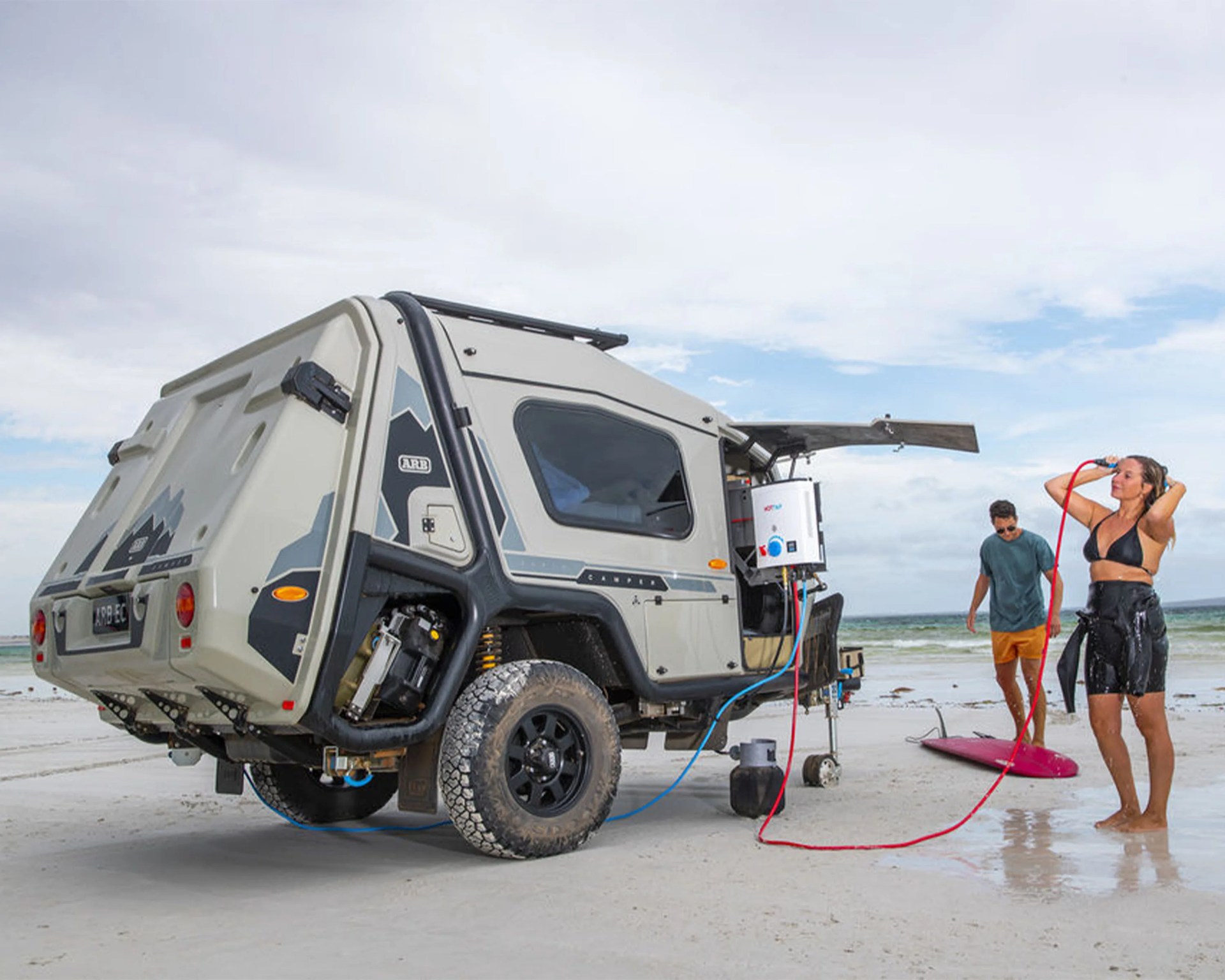Off-road camper trailer with outdoor shower setup on a sandy beach with two people nearby.
