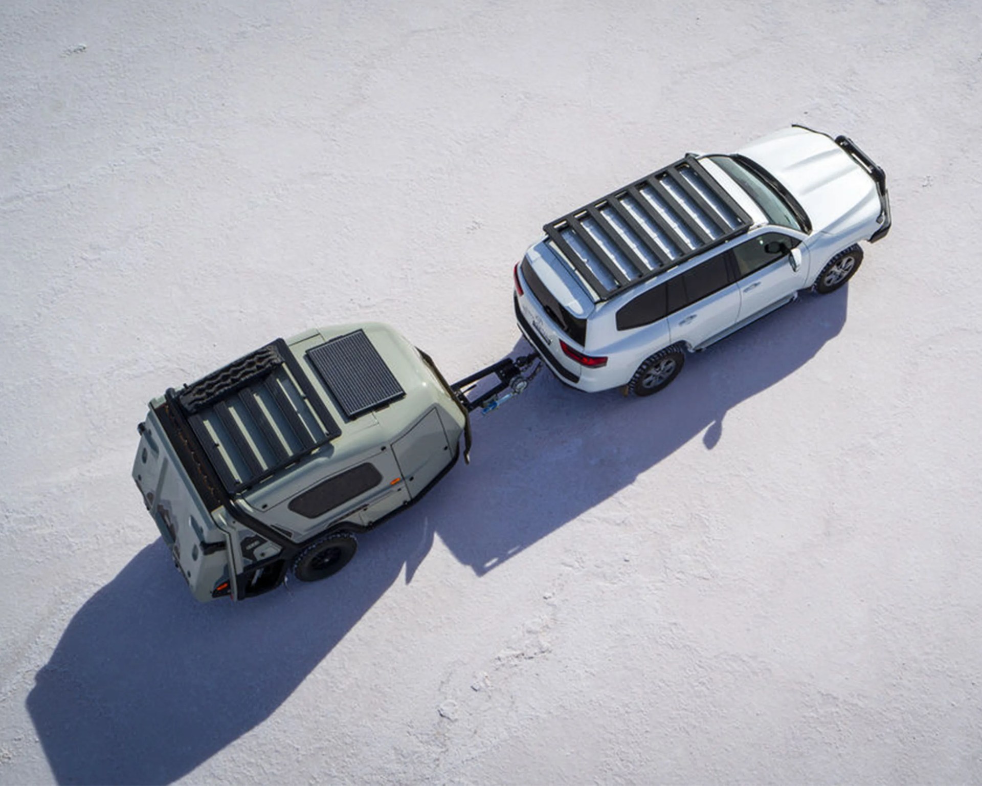 White SUV with roof rack towing a compact, rugged camper trailer with a solar panel on a flat, light-colored surface.