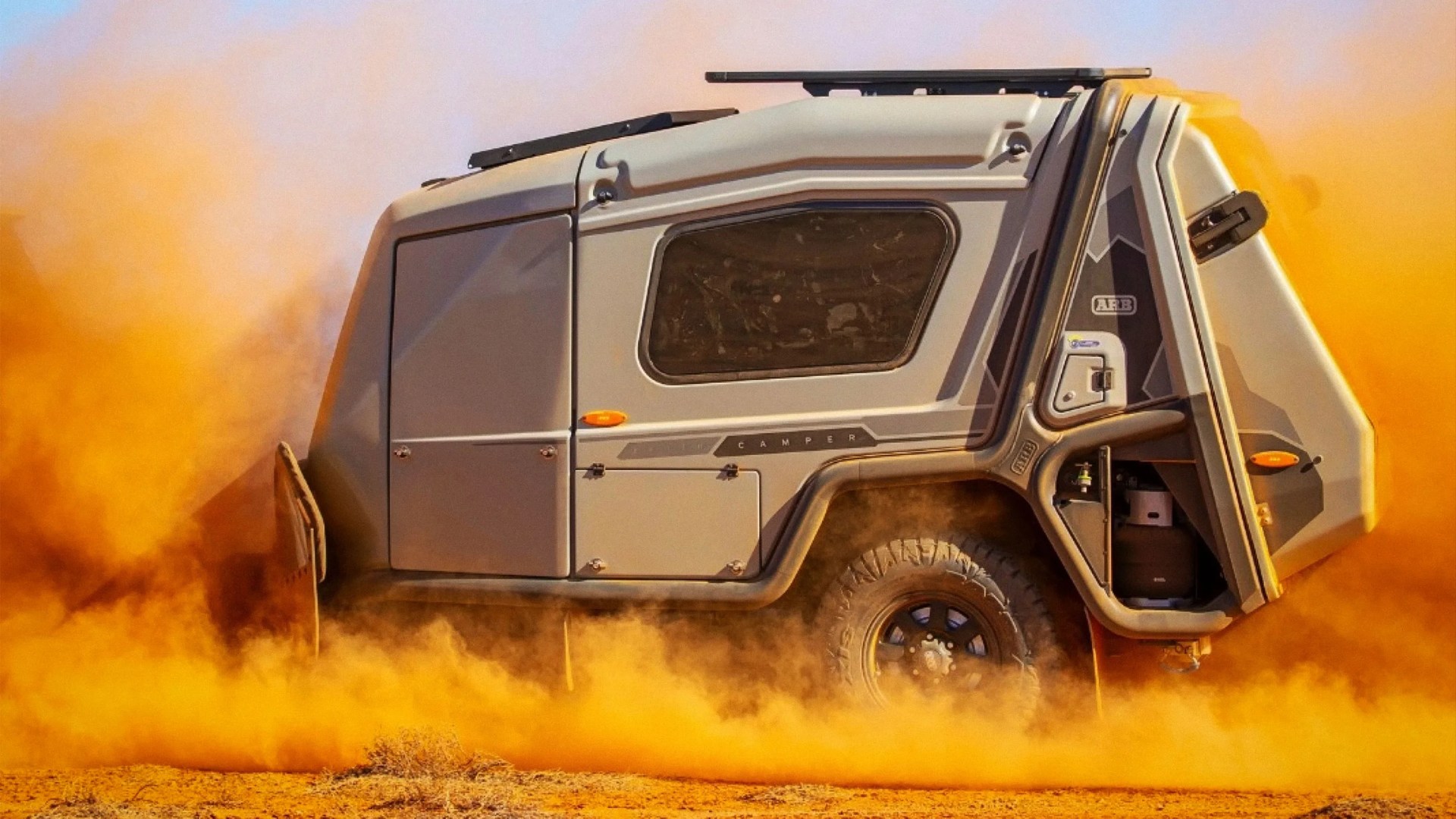 Off-road camper trailer with rugged tires kicking up orange dust in a desert setting.