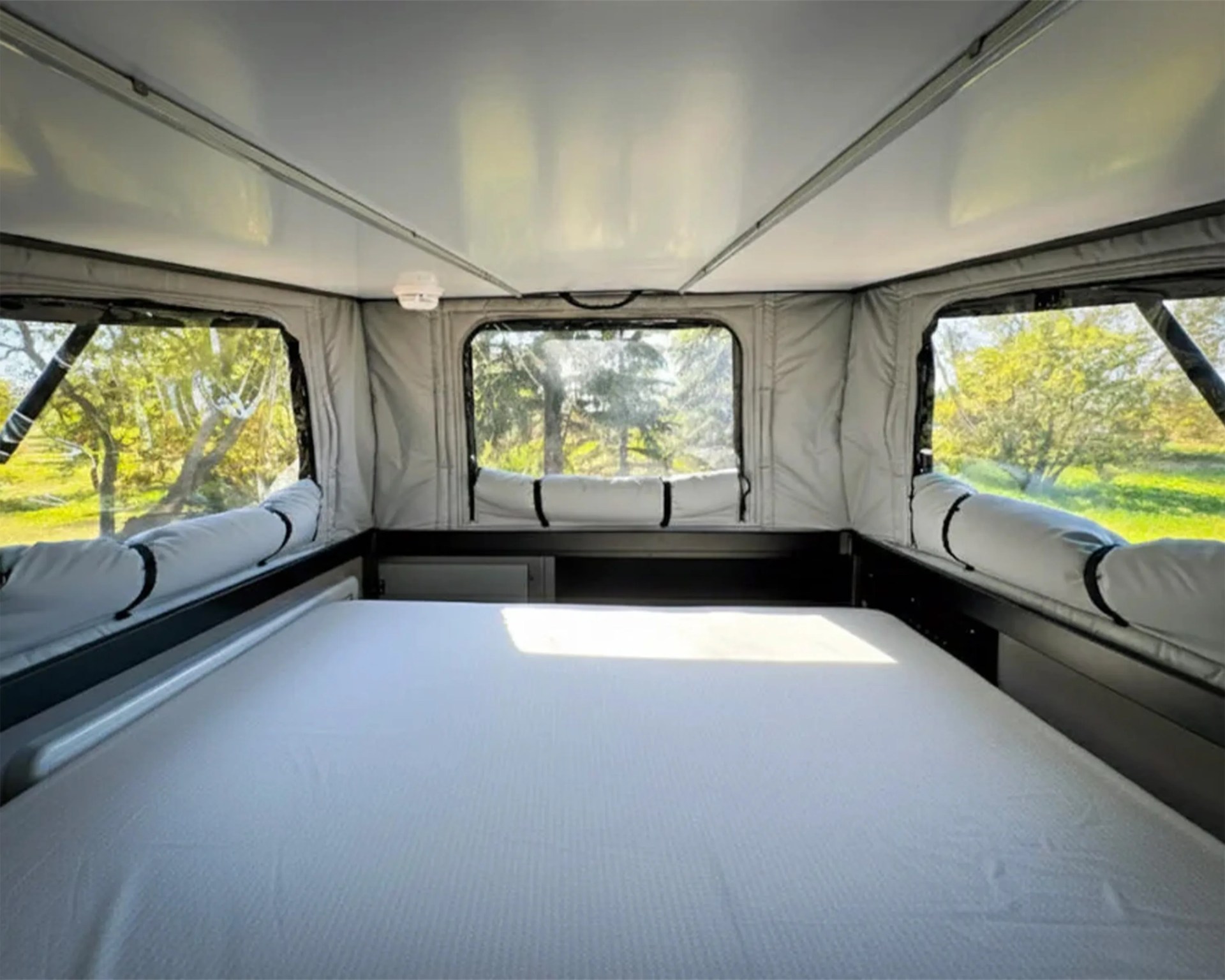 Interior of a camper with a white mattress and large windows showing green trees outside.