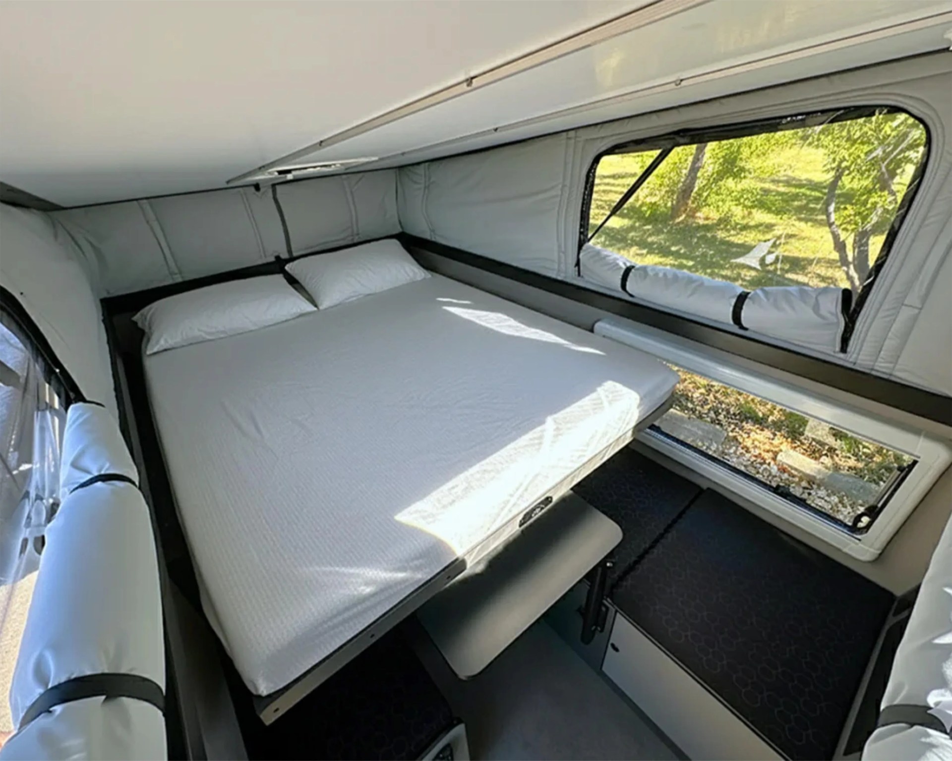Compact camper interior with a raised double bed, two pillows, and large windows showing a green outdoor scene.