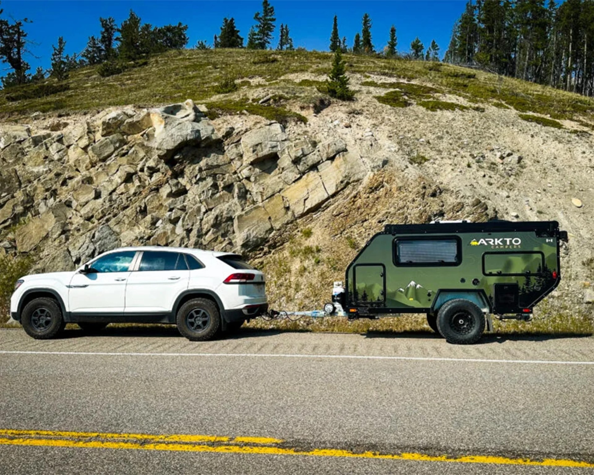 White SUV towing a green Arktō camper trailer parked beside a rocky hillside under a clear blue sky.