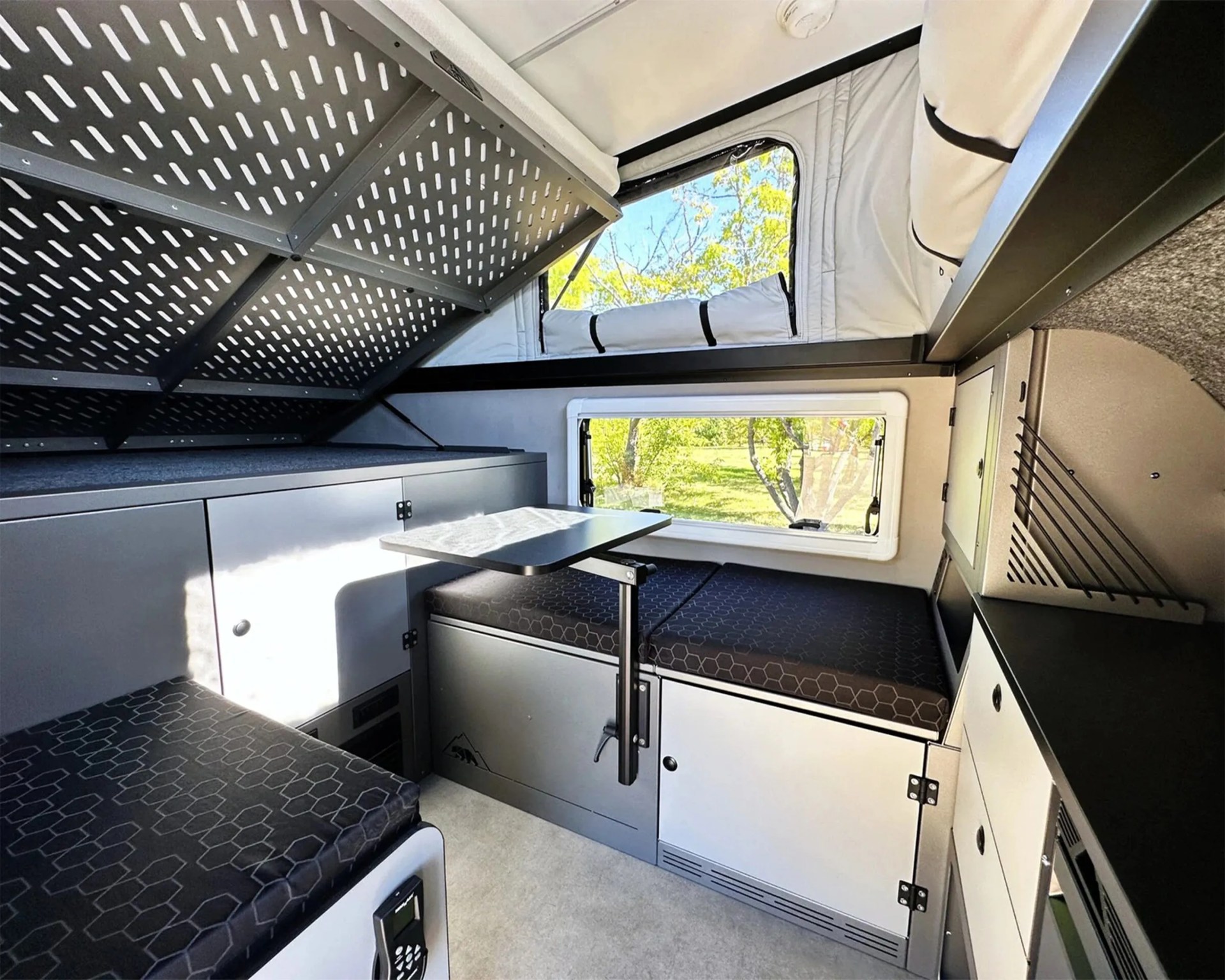 Compact camper interior with black hex-patterned cushions, foldable black table, gray cabinets, and windows showing green trees outside.