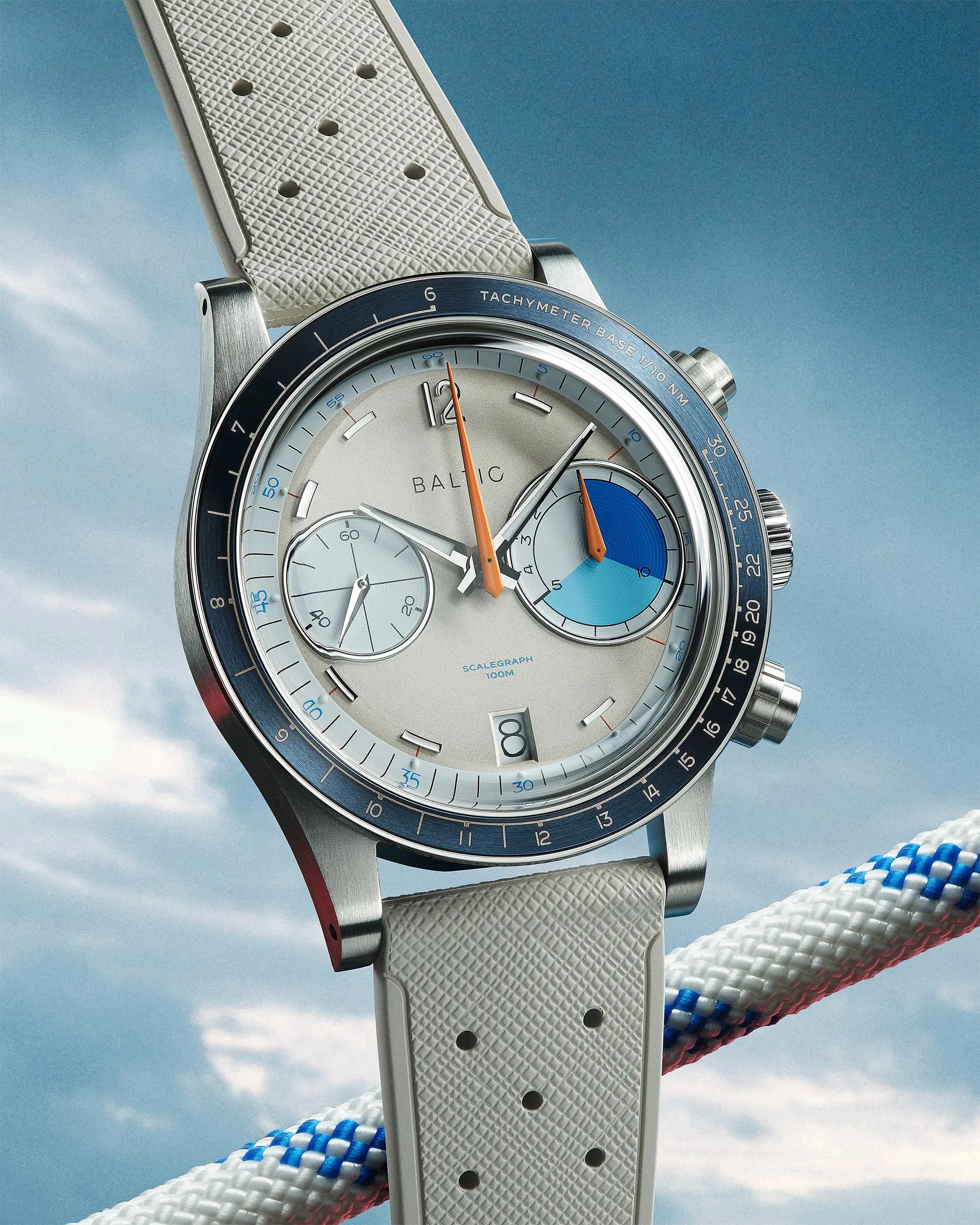 Silver Baltic Scalagraph wristwatch with blue tachymeter bezel and white textured rubber strap against a sky background.