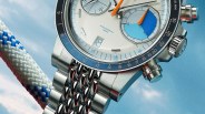 Close-up of a silver chronograph watch with blue and white dial, orange hands, and a metal link bracelet.