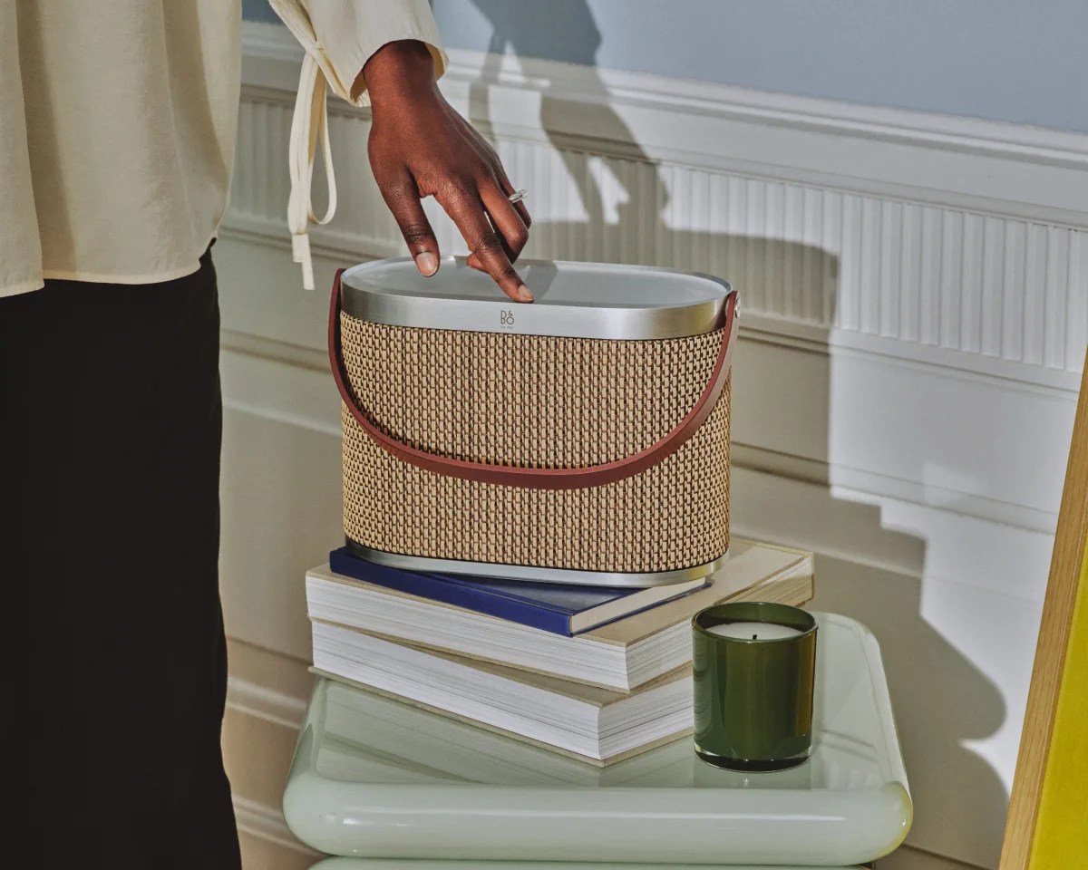 Portable woven speaker with leather strap and silver top, placed on stacked books on a glass table.
