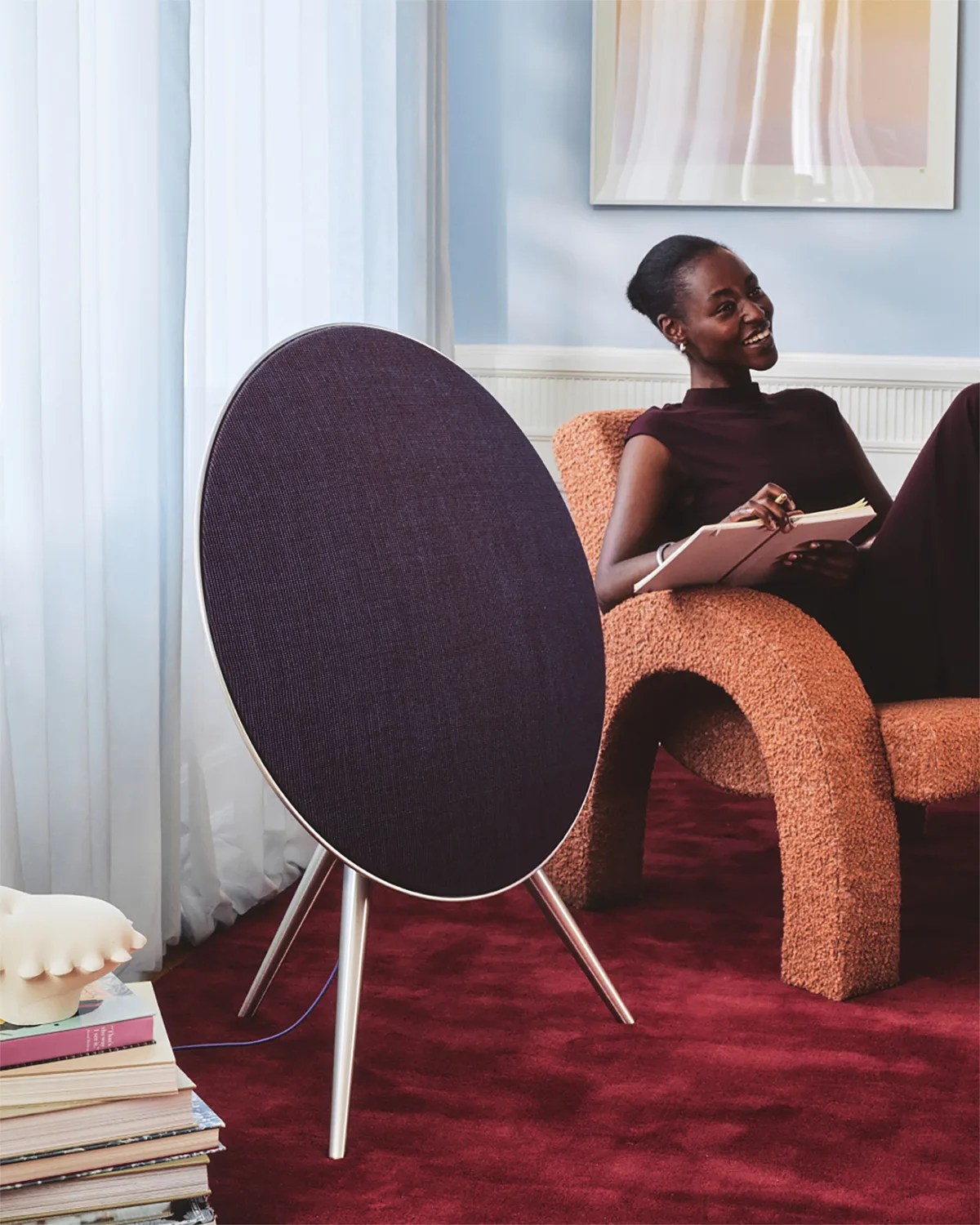 Large round black fabric speaker on silver tripod legs on a red carpet next to a person sitting on a textured orange chair.