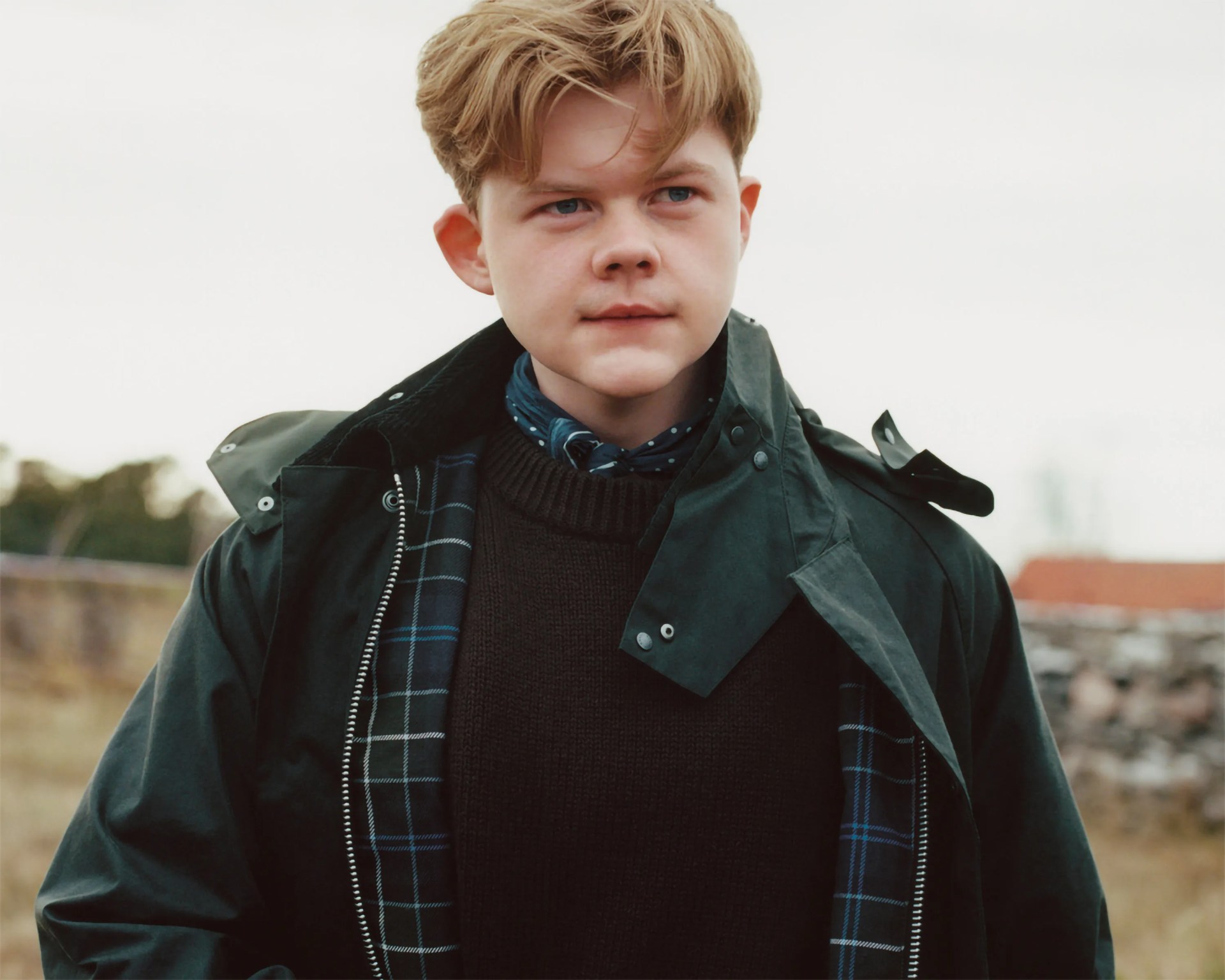 Young person with short light brown hair wearing a dark green jacket over a black sweater and a blue patterned scarf.