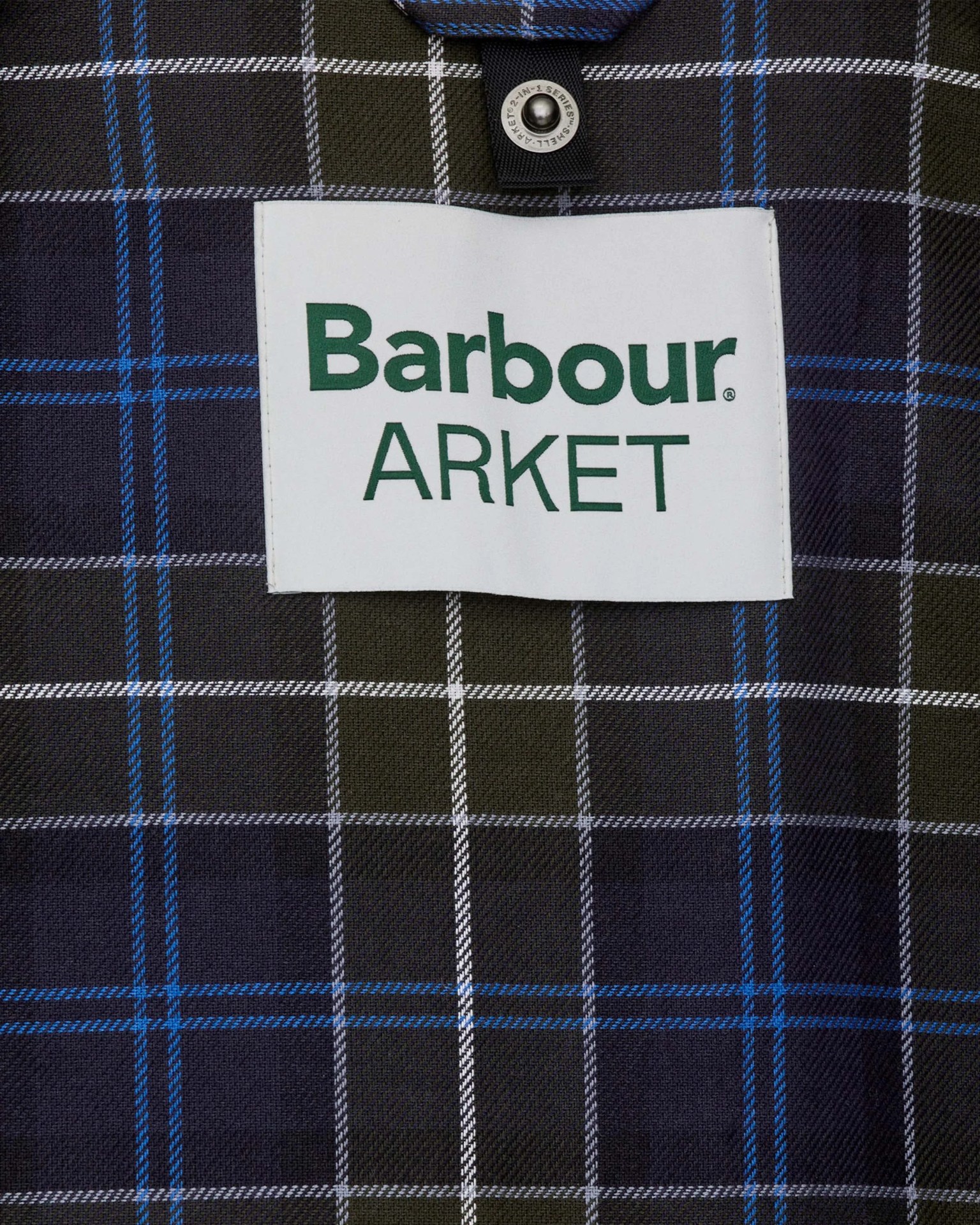 White fabric label with green "Barbour ARKET" text on dark plaid fabric with blue and white lines.