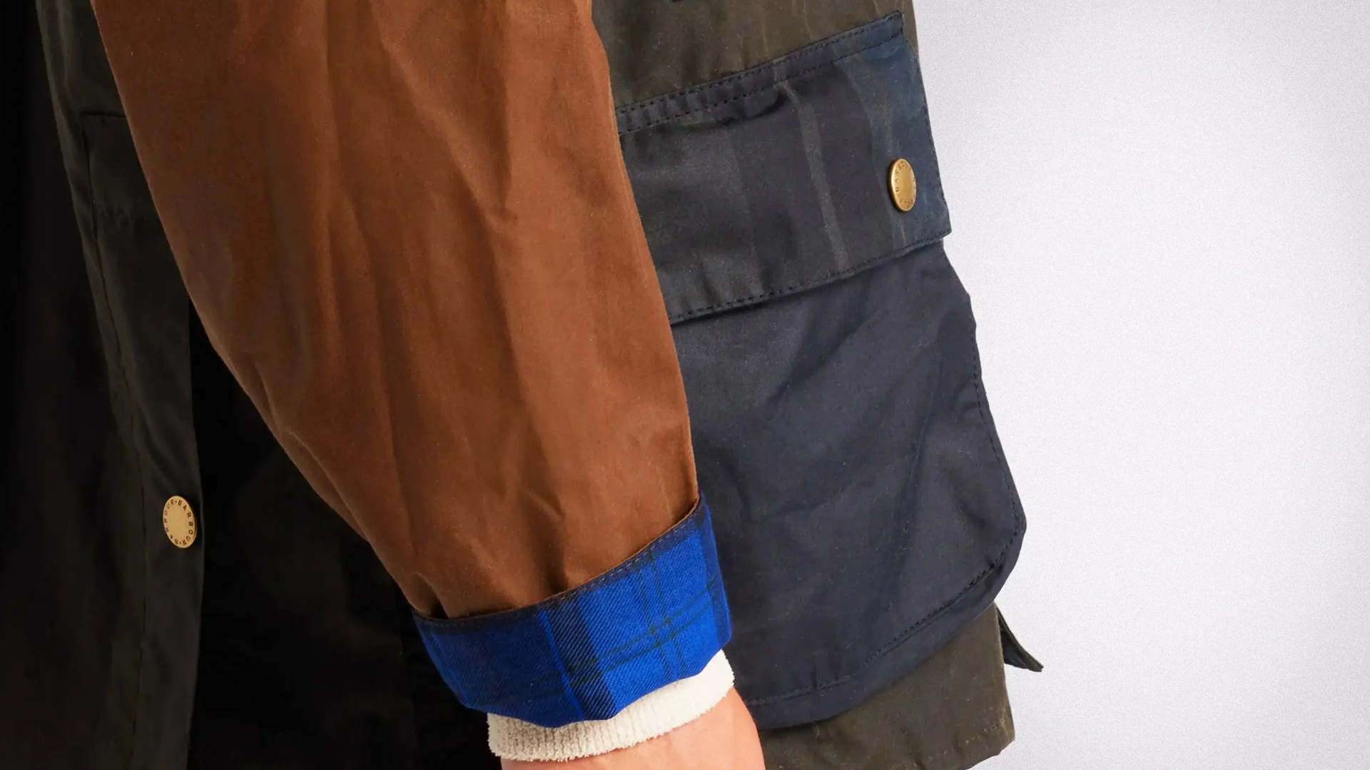 Close-up of a jacket sleeve with brown fabric and blue plaid cuff over a white knit wristband, next to a navy pocket with a brass button.