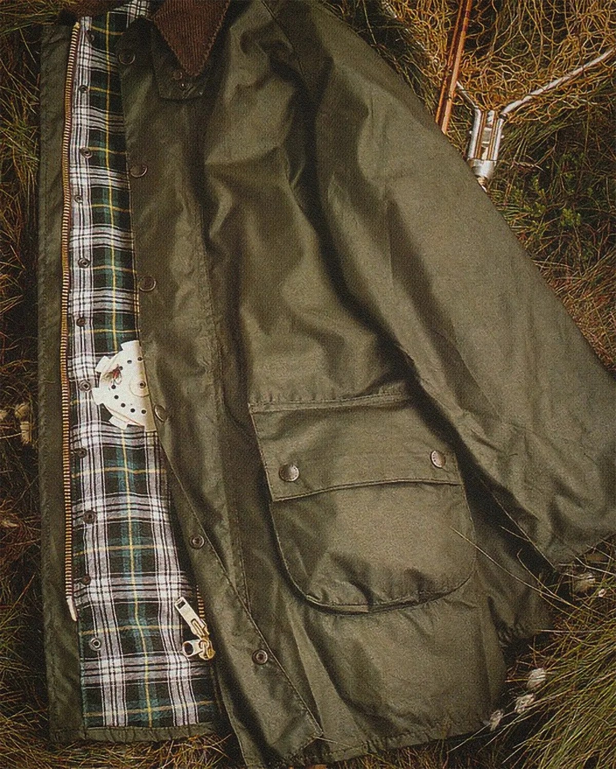 Olive green waxed cotton jacket with plaid lining and snap button pockets laid on grass.