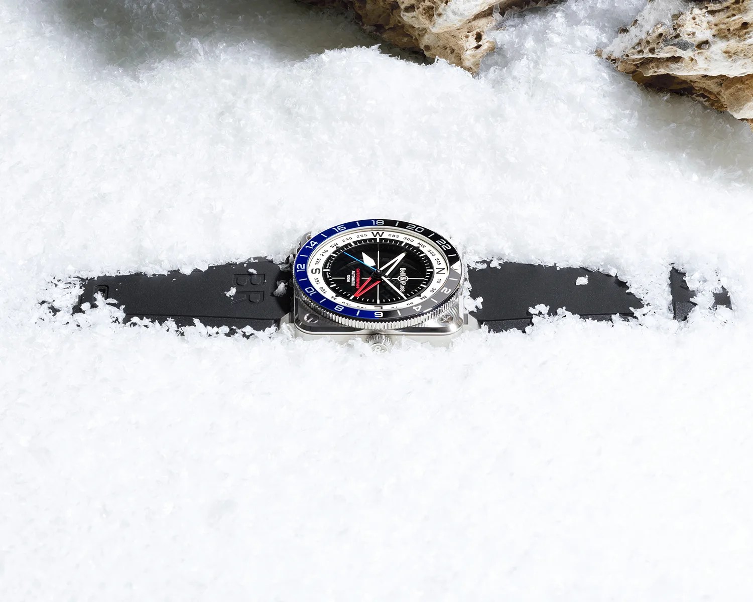 Square silver watch with black dial, blue and black bezel, and black rubber strap partially buried in snow.