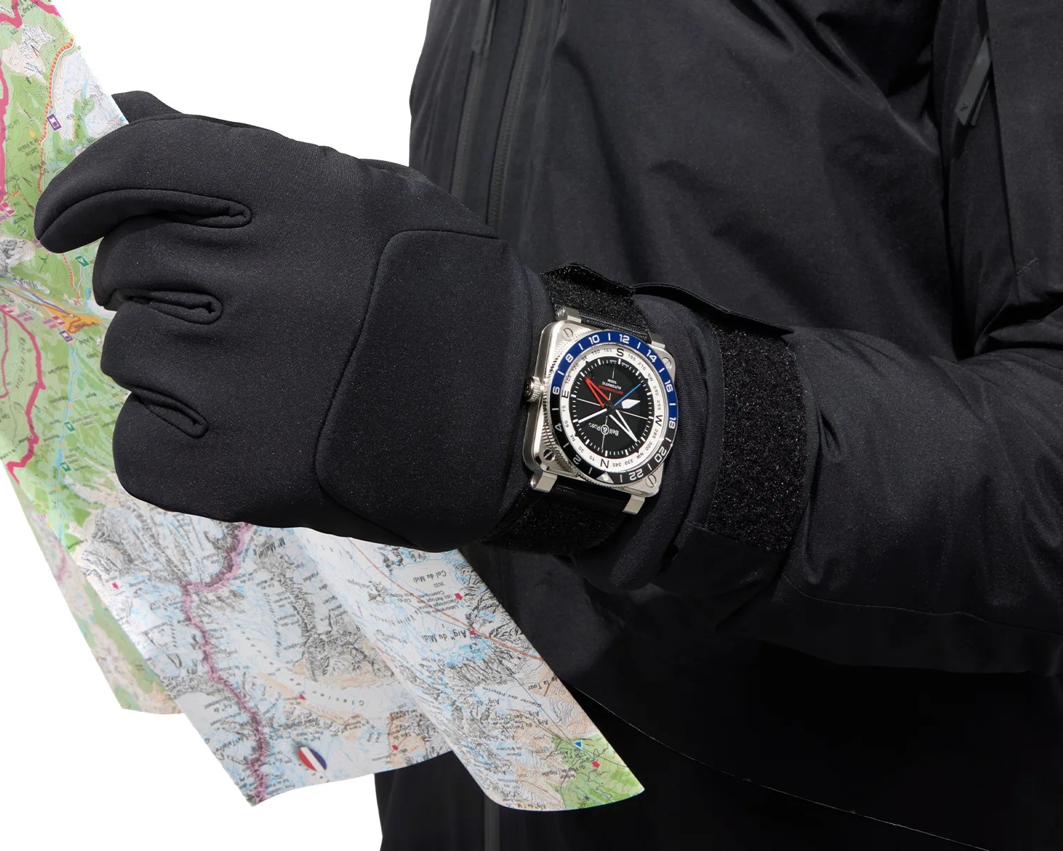 Black gloved hand holding a folded map, wearing a square silver watch with a black dial and blue and white bezel.