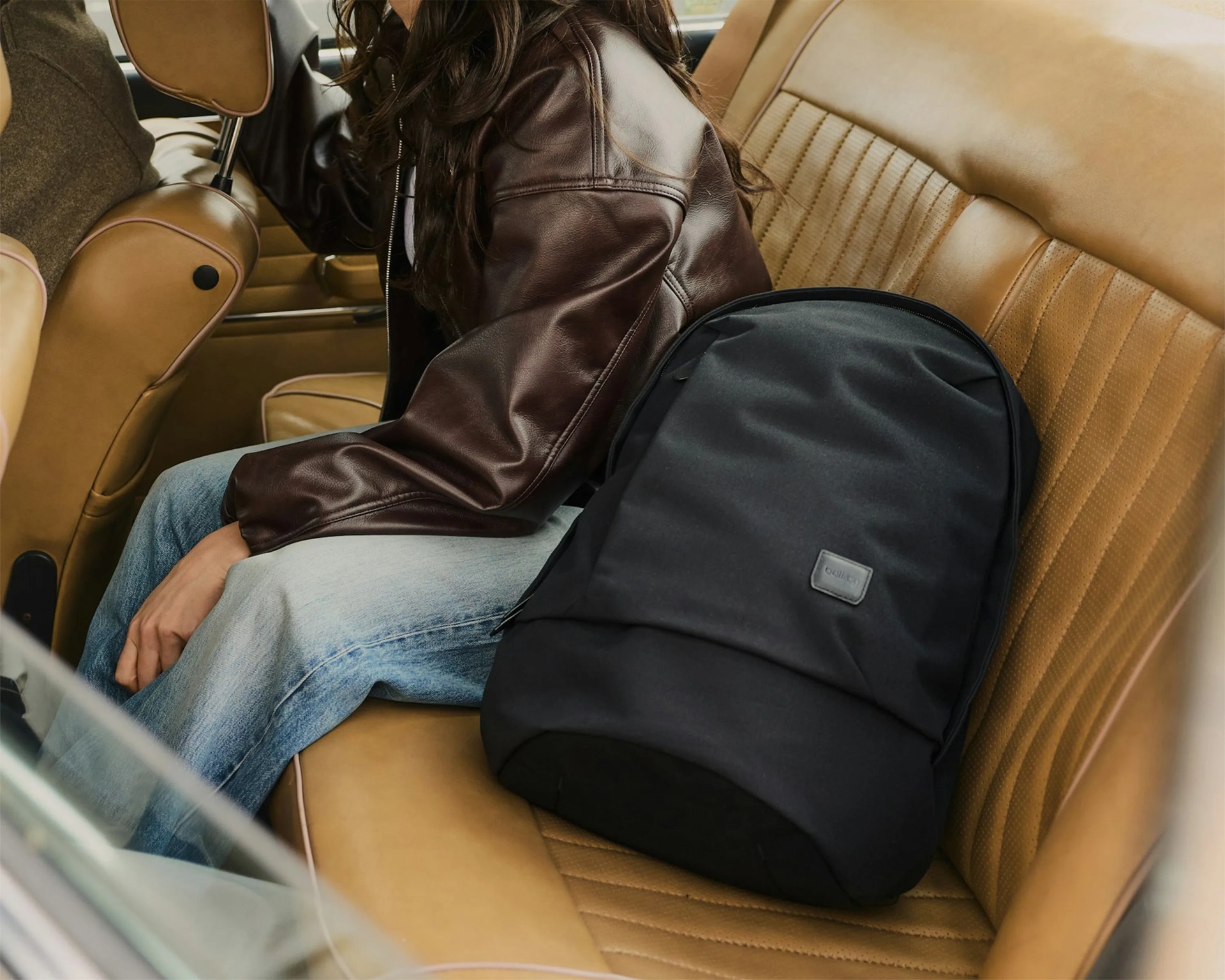 Black fabric backpack with a small logo patch on the backseat of a tan leather car interior next to a person wearing a brown leather jacket and blue jeans.